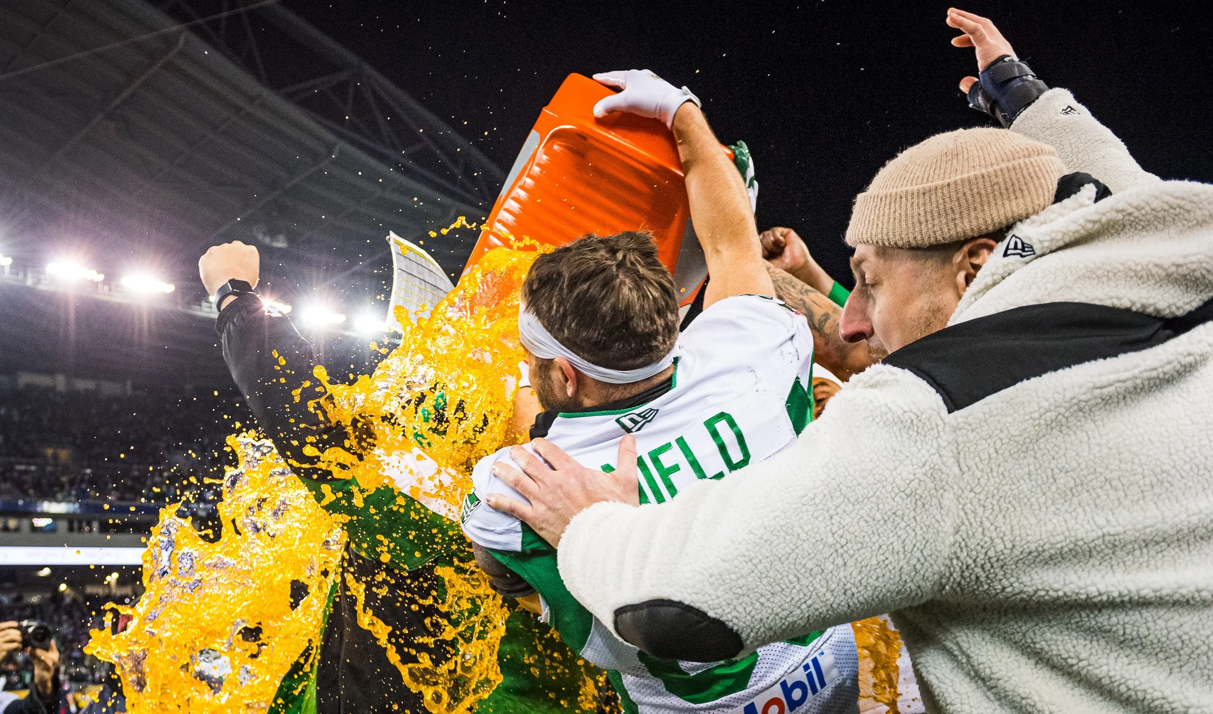 Athletes celebrating with a beer dump celebration on the field at night, with stadium lights and a crowd in the background.