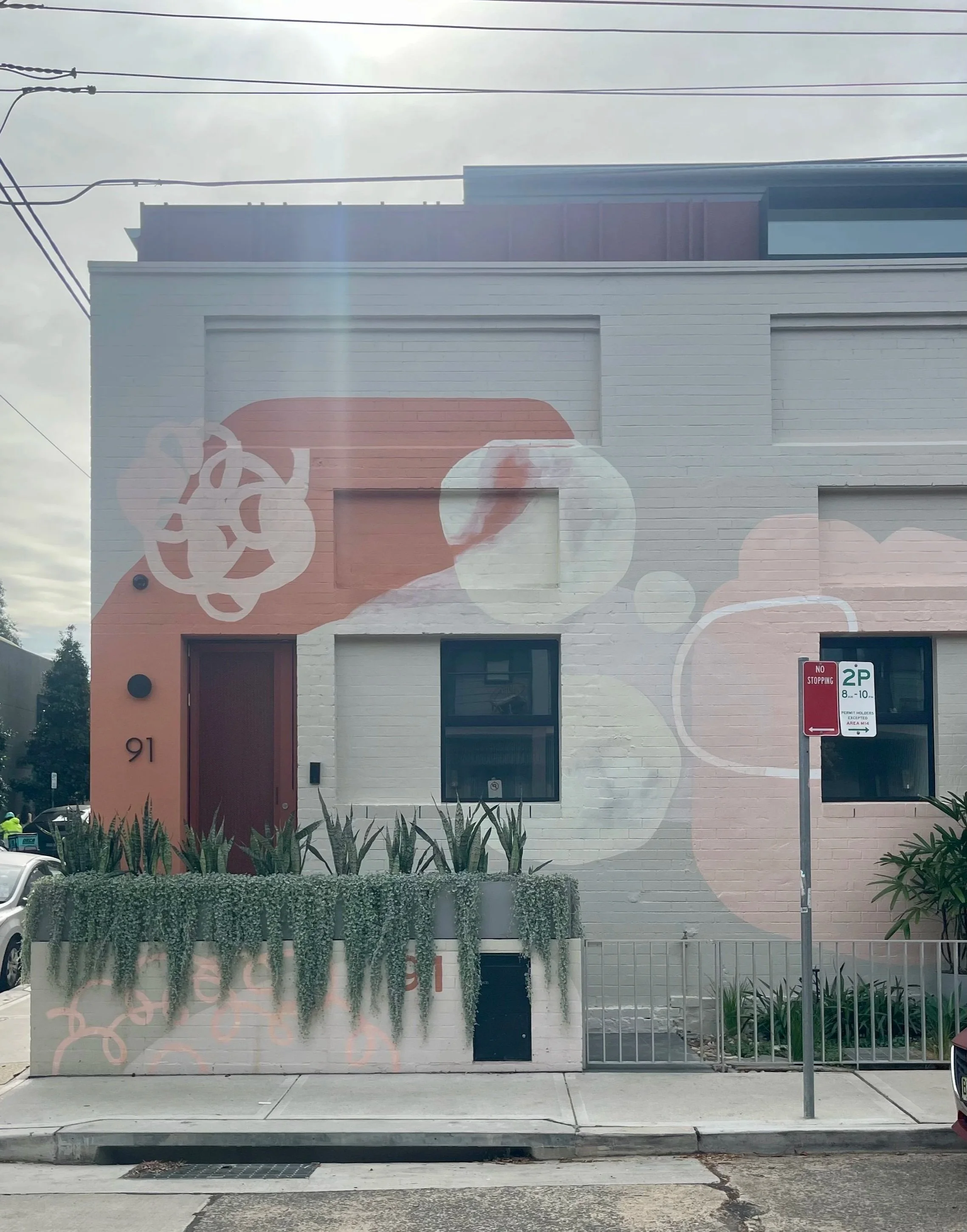 Photo of a building with a painted mural of a woman with glasses and a bun, surrounded by abstract shapes. The building has a white brick exterior, a black door, and several large windows. There are a few plants in front and a parking sign on the sidewalk.
