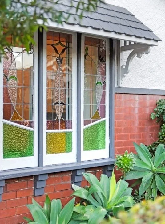 A close-up view of a stained glass window on a brick house, surrounded by green plants. Home is a renovation located in Marrickville.