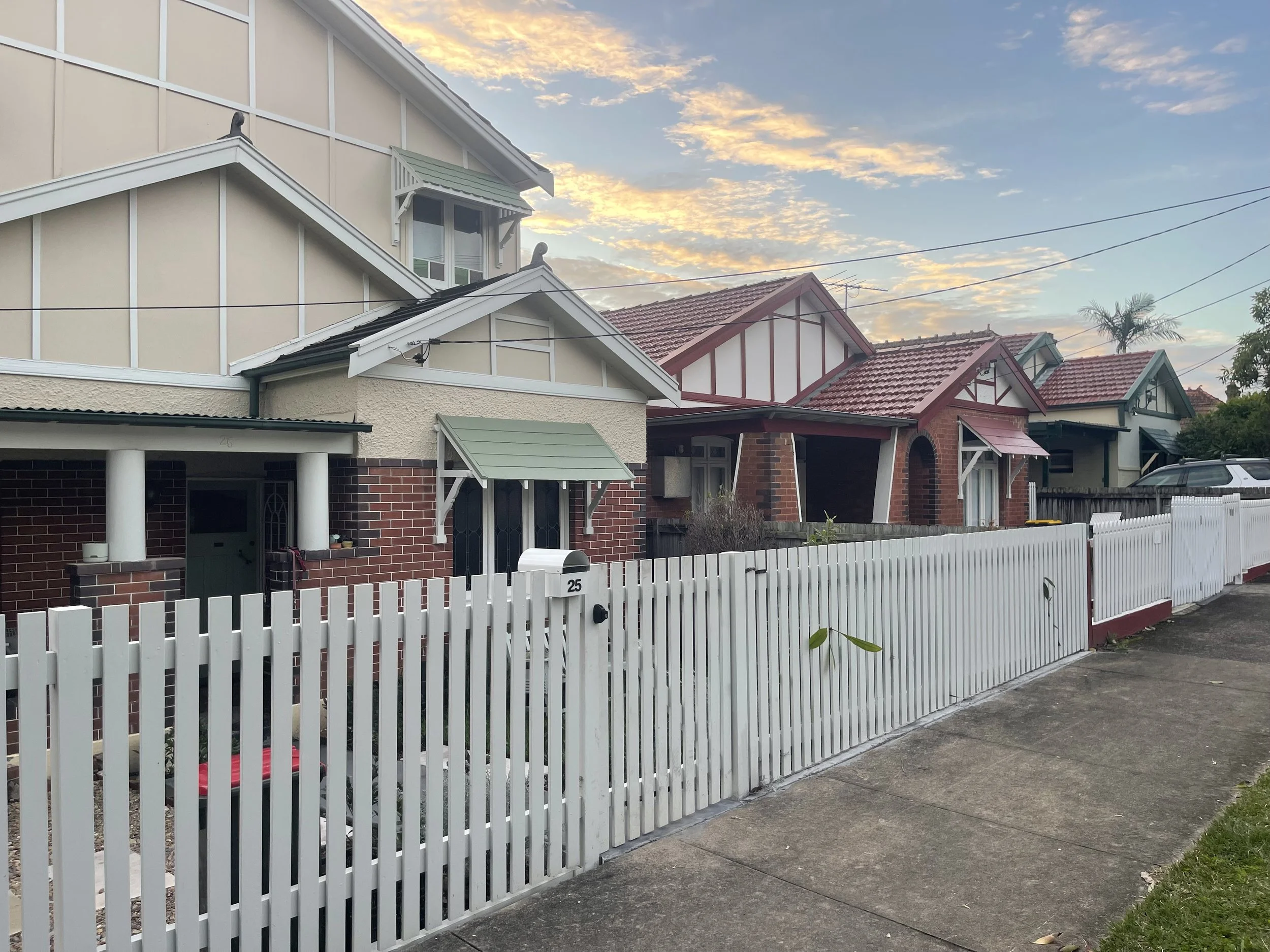 A row of suburban houses with various architectural styles and colors, with a white picket fence in front, and a partly cloudy sky at sunset in the background.