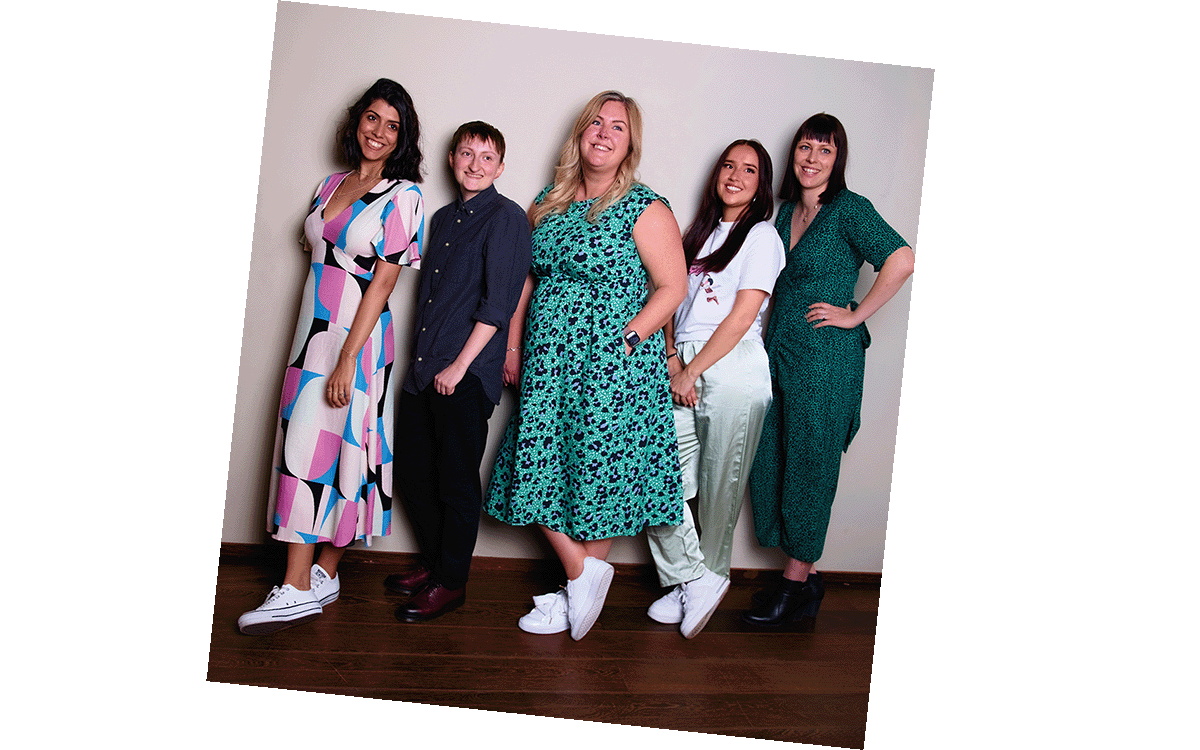 Group of six women standing against a plain wall, smiling at the camera in a casual setting.
