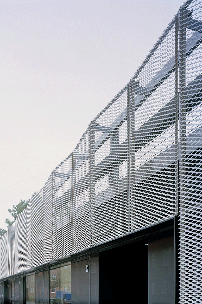 Das gestalterische Konzept des 102m langen Gebäudes mit Streckmetallfassade versteht sich einerseits als guter Nachbar zur Wohnbebauung an der Scharnhorststraße und andererseits als selbstbewusste und attraktive Eingangssituation für den Campus. Durch die verwendeten Materialien und Oberflächen entsteht ein lebendiger und anregender Dialog zwischen dem neuen Gebäude und den bestehenden ehemaligen Kasernengebäuden auf dem Campus. In der äußeren Gestaltung erscheint das Gesamtobjekt mit der verwendeten Streckmetallfassade gleichzeitig einheitlich und vielseitig. Die Fassade fasst das Gebäude mit seinen verschiedenen Nutzungen zu einem Baukörper zusammen. Dennoch wirkt sie in ihrer Textur abhängig vom Standpunkt des Betrachters sowie vom Sonnenstand im Tages- und Jahresverlauf transparent und abwechslungsreich. Das Streckmetall ist weiß glänzend beschichtet und erzeugt einen bewussten Kontrast zu den umgebenden Ziegelfassaden. Es umhüllt den Baukörper attraktiv und elegant und zeigt den Charakter eines technischen Gebäudes auf dem Campus.