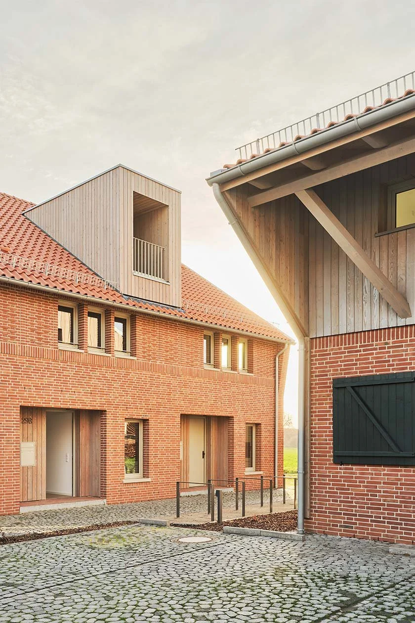  Das zur Straße orientierte Gebäude mit vier Wohneinheiten nimmt die Form einer klassischen Remise auf. Es ist zurückversetzt positioniert und giebelständig ausgerichtet. Die klare Kubatur und die prägnante Dachform zitieren den Bautypus landwirtscha