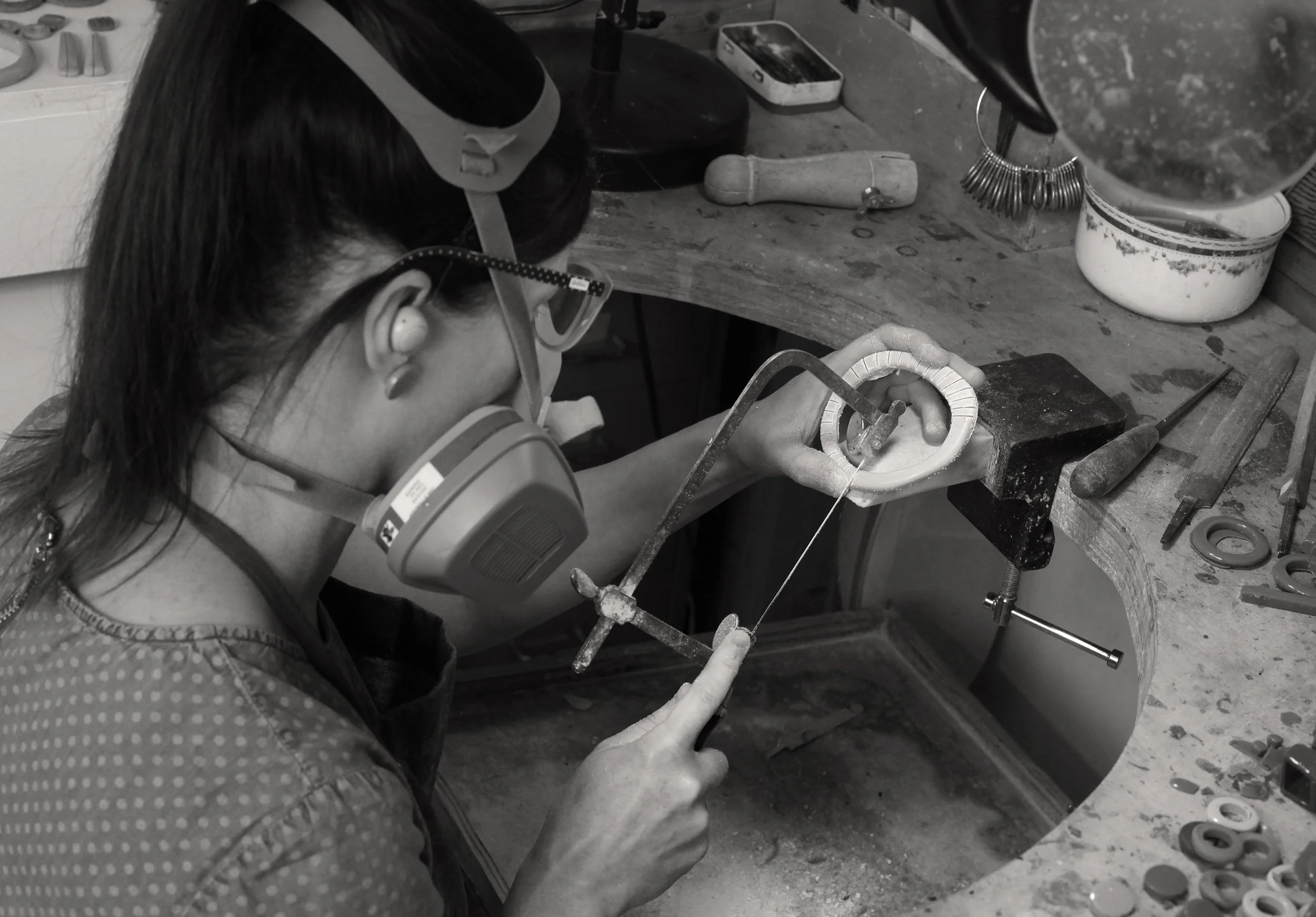 A woman wearing a face mask and goggles, working on a piece of resin jewellery at a jewellery bench, with a saw and other tools in a workshop.