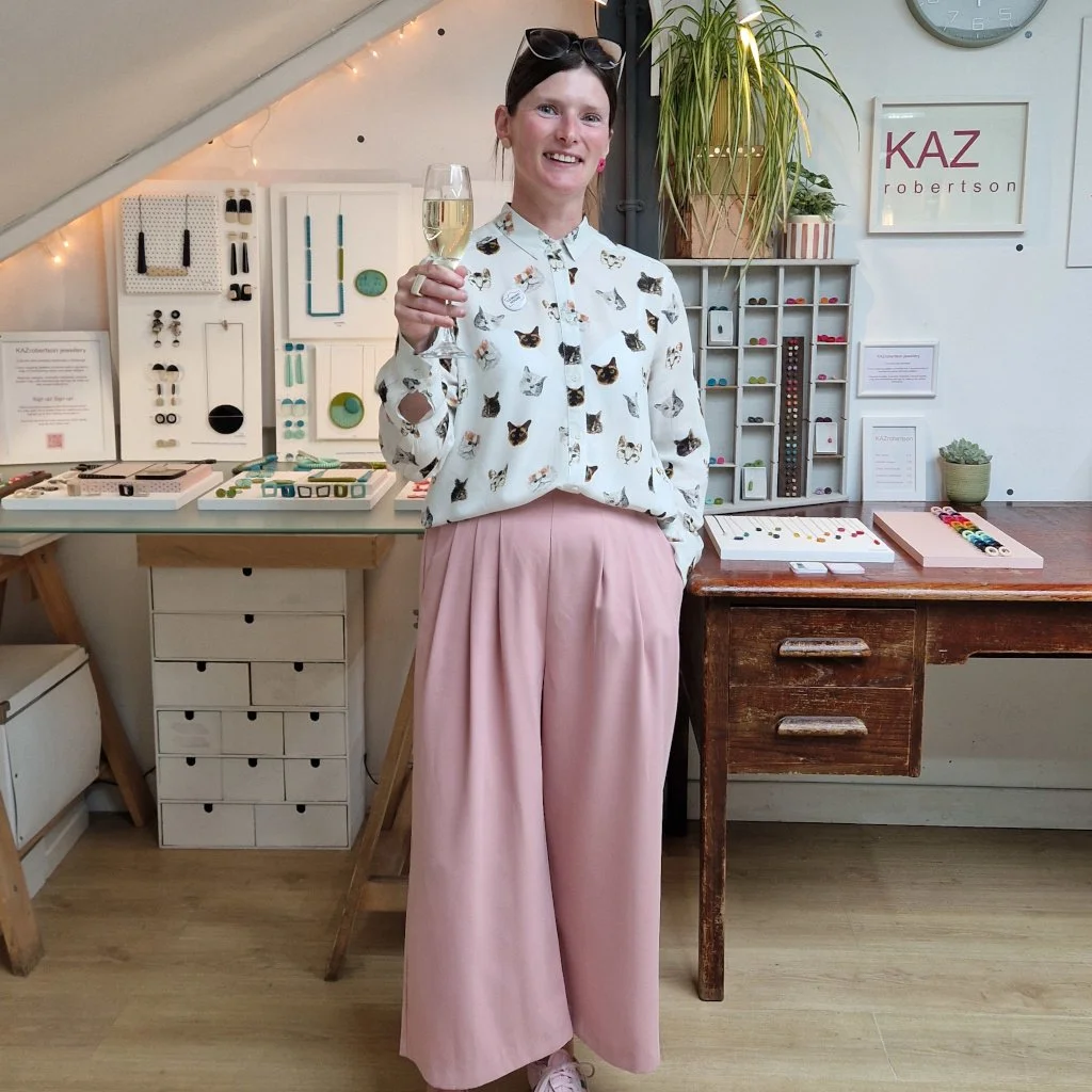 Stylishly dressed woman in cat shirt and pink trousers in front of her resin jewellery display