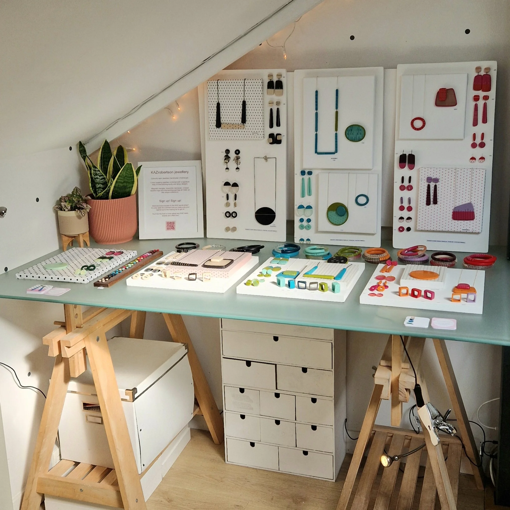 Display of colourful jewellery including earrings, bangles, and rings arranged on white panels and trays, with a plant in a pink pot and a sign-up sheet on a table.