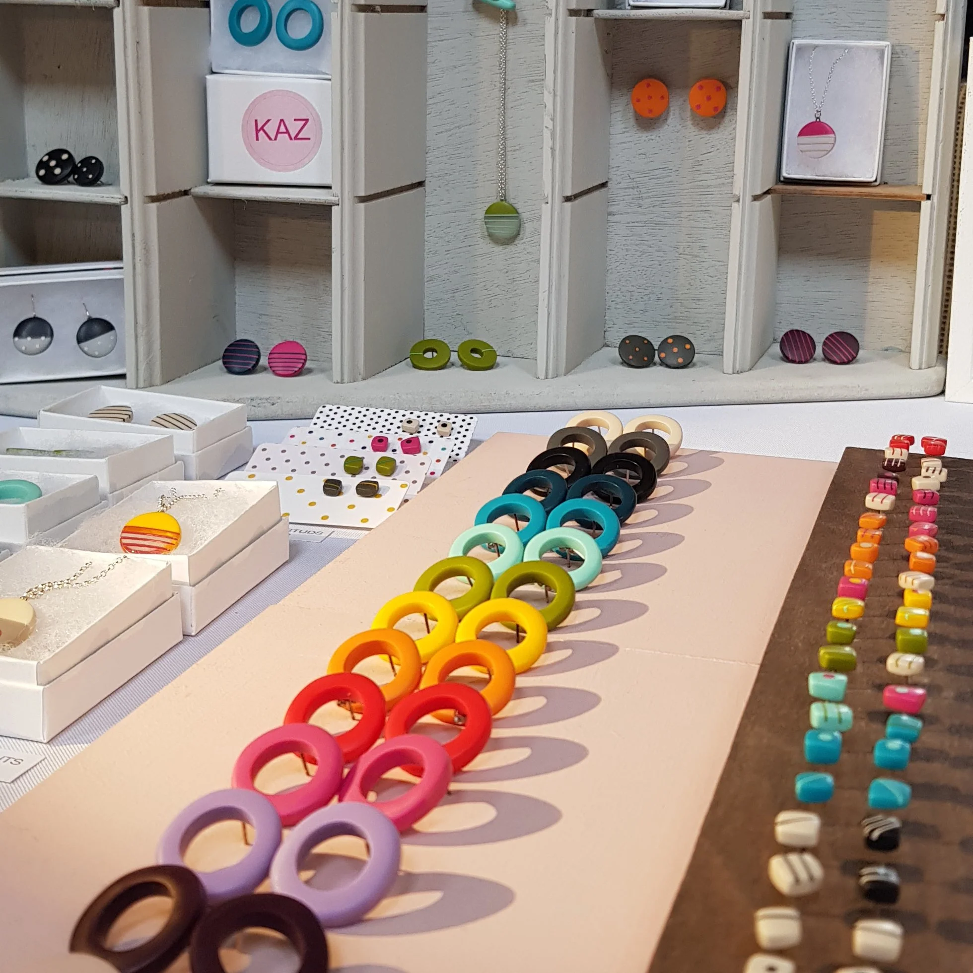 A display of colourful resin jewellery, tiny stud earrings, hoop earrings in a rainbow line display. Some small jewellery pendants and studs boxed as gifts in the background