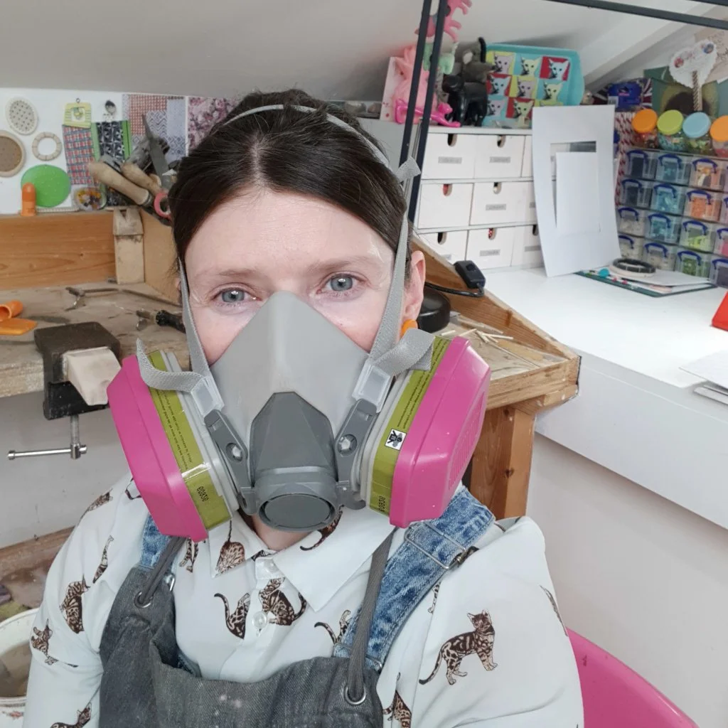 A girl with blue eyes and brown hair wearing a full protective mask with pink filters, a white shirt with a big cat pattern, and an apron, in a jewellery studio space surrounded by tools and supplies.