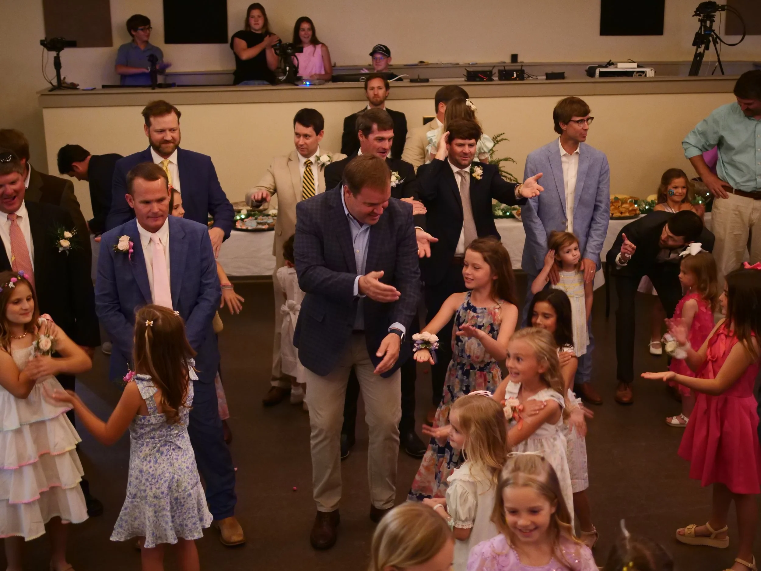 Group of adults and children dancing at a celebration or party, with a table of food and drinks in the background.
