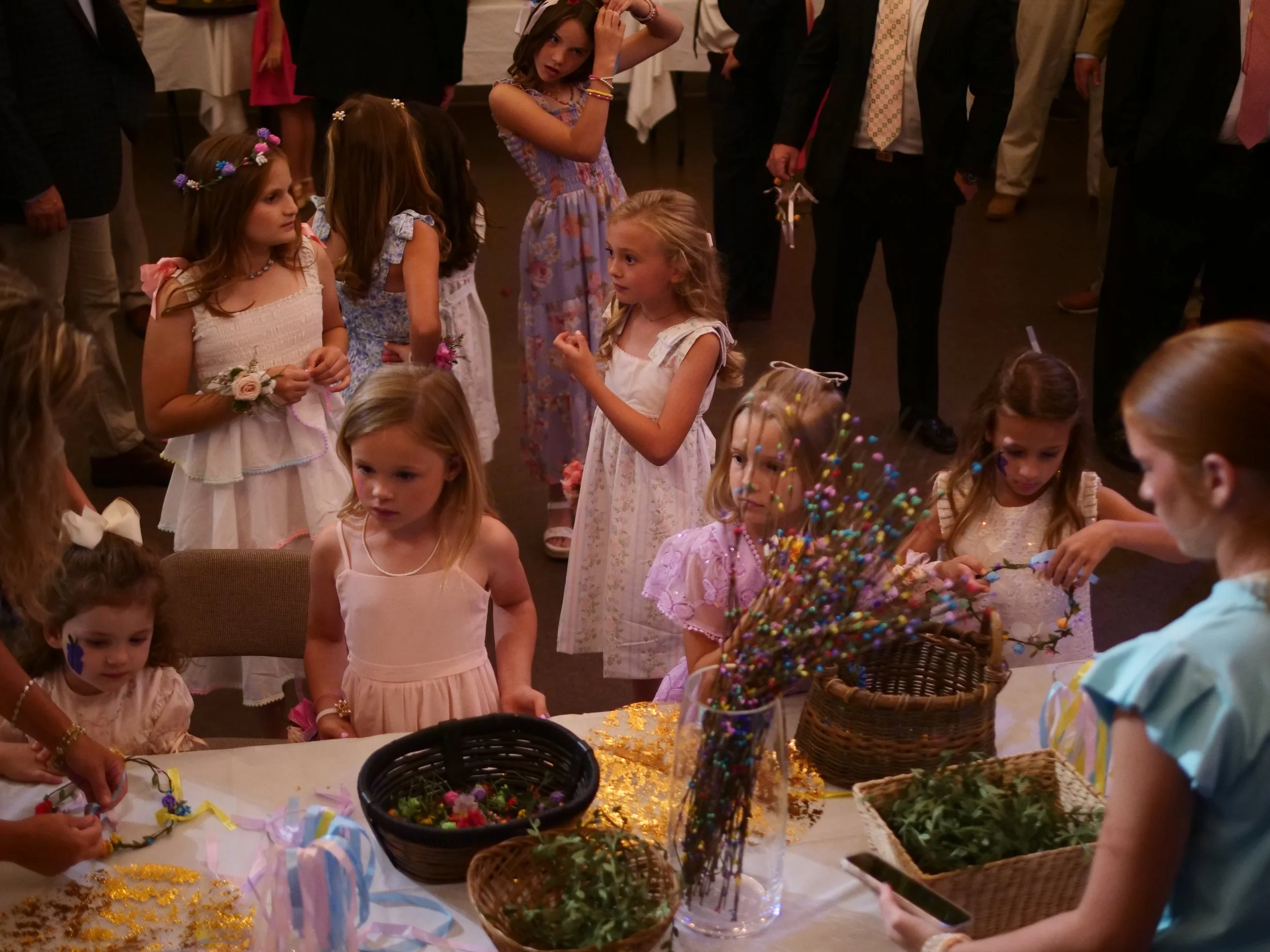 Children celebrating, wearing dresses, surrounded by women and men in formal attire, with tables decorated, some with colorful ribbons, glitter, and baskets of flowers or plants, likely at a party or festive event.