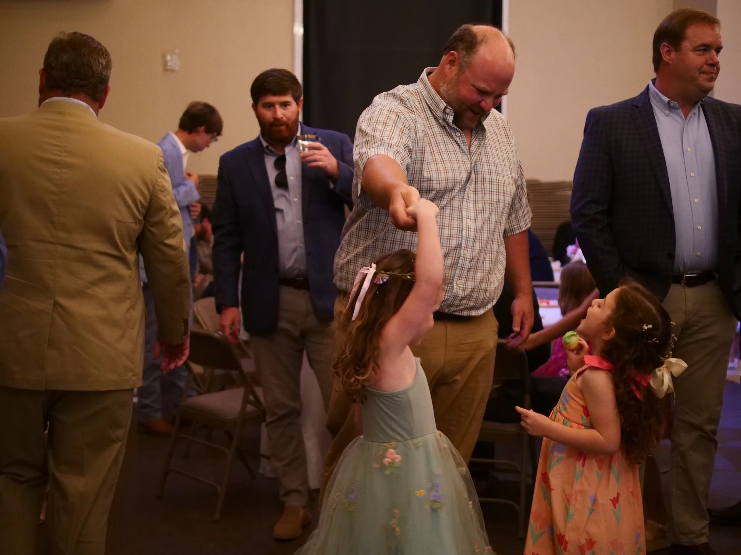 A group of adults and children gathered indoors, engaging in a social activity. The children are wearing colorful dresses, and one girl is holding a flower. The adults are dressed in casual and business attire, smiling and interacting with the children.