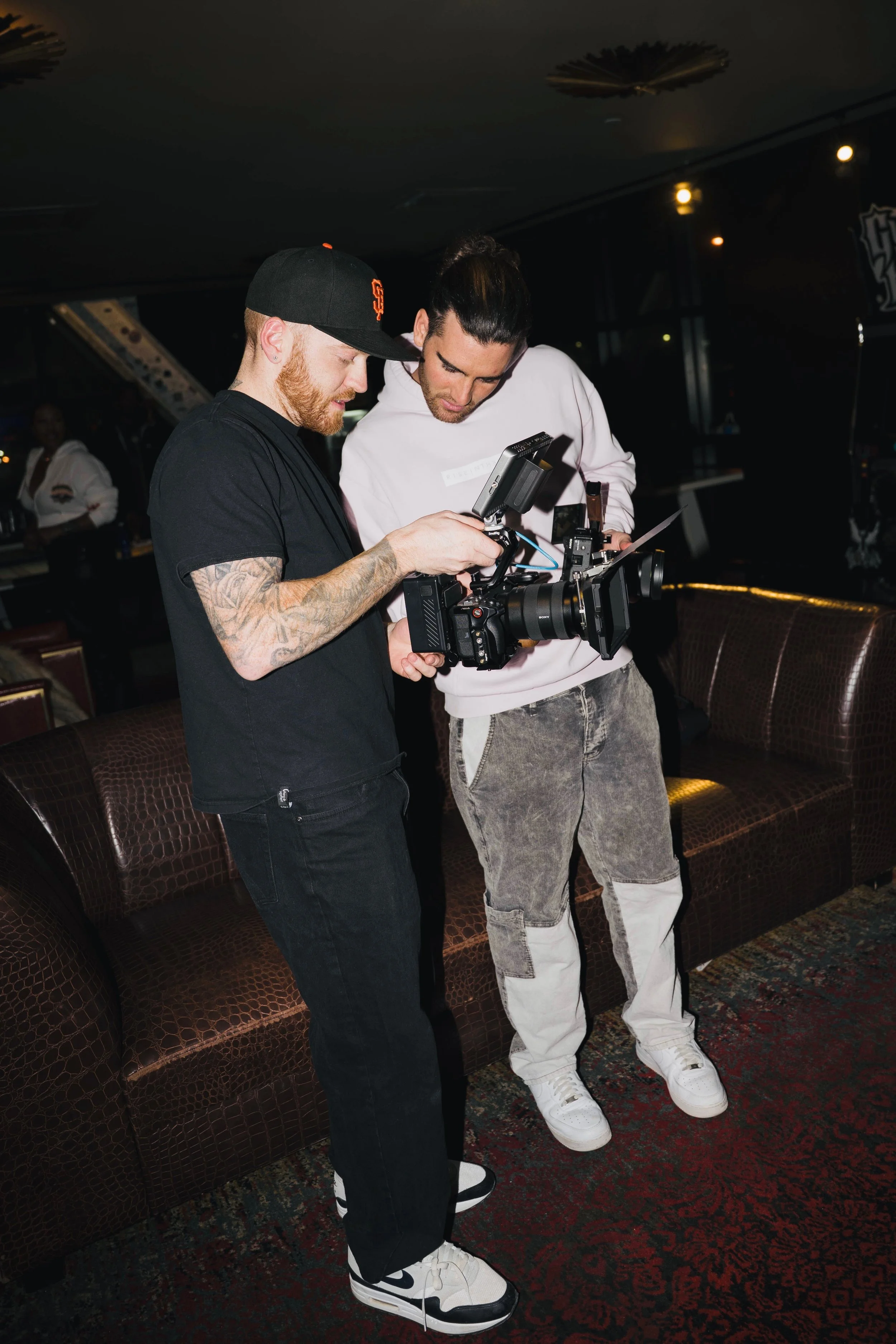 Two men are looking at a professional camera while standing on a patterned carpeted floor in a dimly lit lounge or bar area. One man has a tattooed arm and is wearing a black cap, black t-shirt, and black pants, while the other man has long dark hair tied back, is wearing a white hoodie, and distressed jeans. There are brown leather couches in the background.