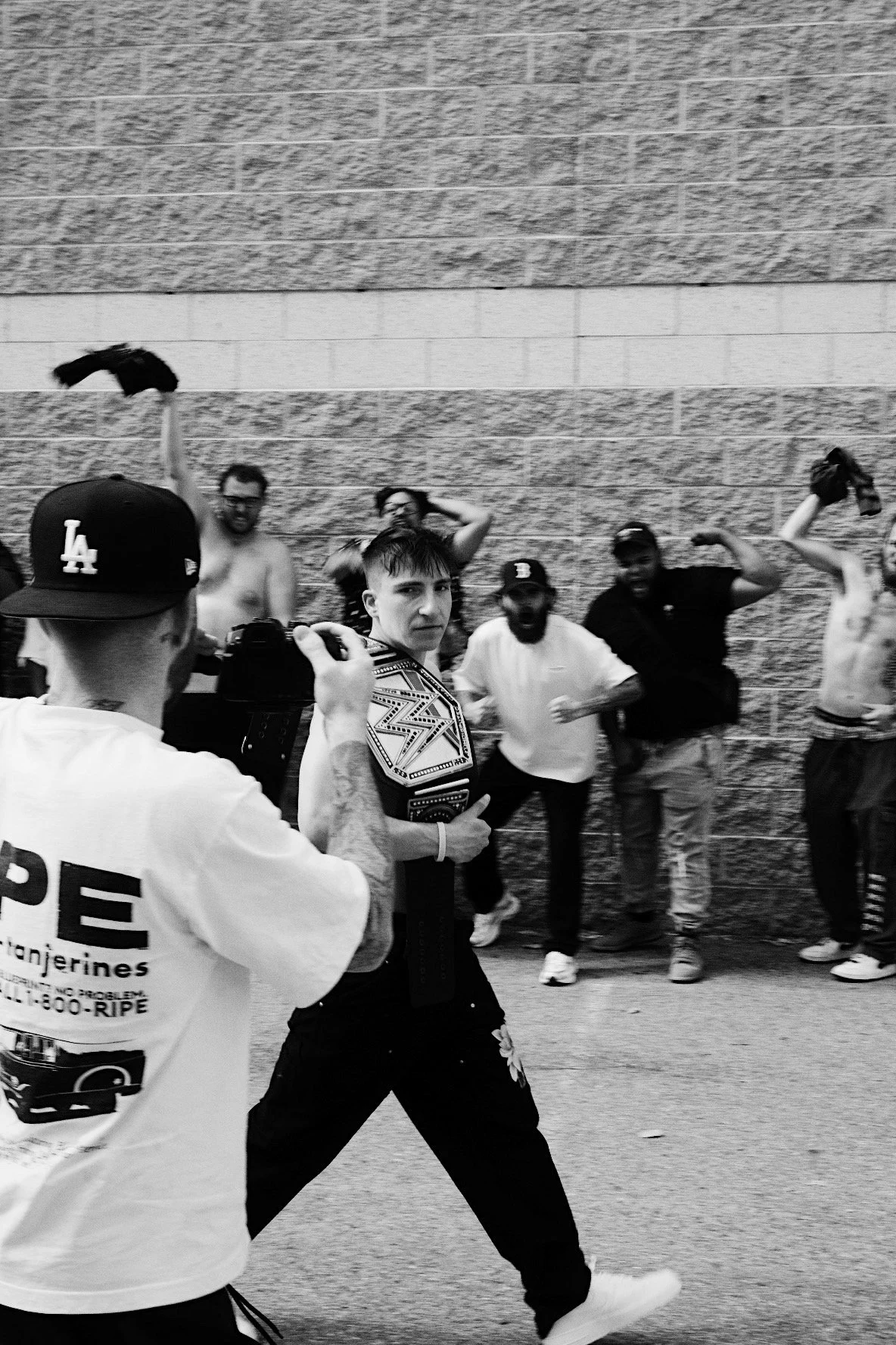 Group of young men, some shirtless, standing against a brick and concrete wall. One man in the foreground is holding a camera and appears to be filming or taking a picture of another man wearing a wrestling belt, who is looking at the camera. The men in the background are posing casually, some with arms raised or resting on their heads.