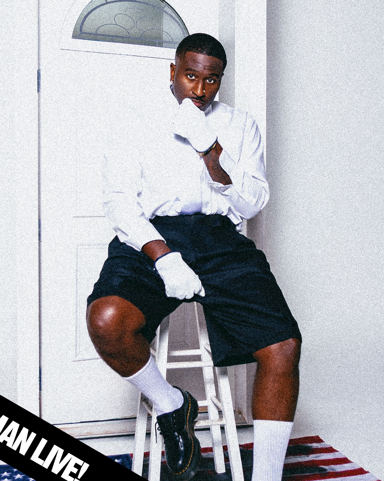 A young man in a white shirt, black shorts, white socks, and black shoes sitting on a bar stool in a room with white walls, a door with an oval window, and an American flag rug. He is wearing white gloves and looking directly at the camera with a serious expression, resting his chin on one gloved hand.