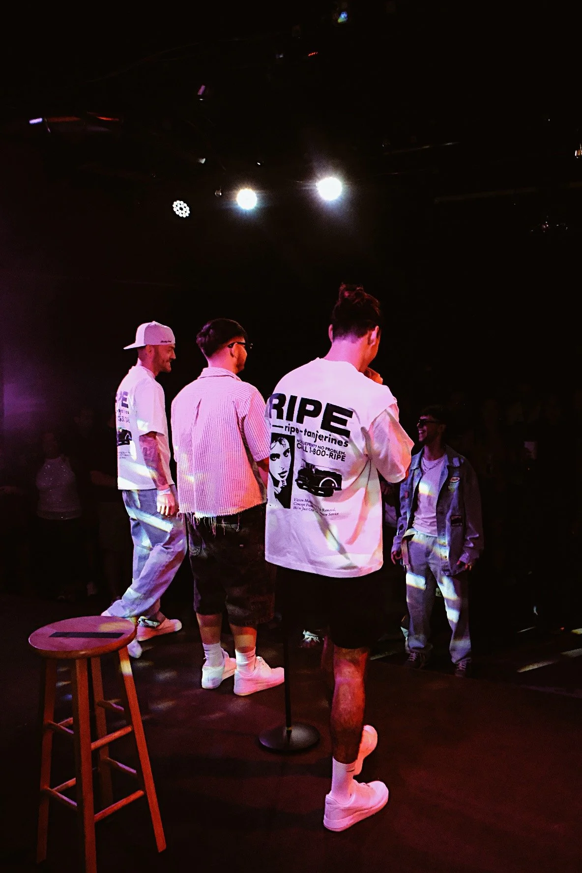 A group of four young men standing on stage under bright lights in a dark room, wearing casual streetwear and sneakers.