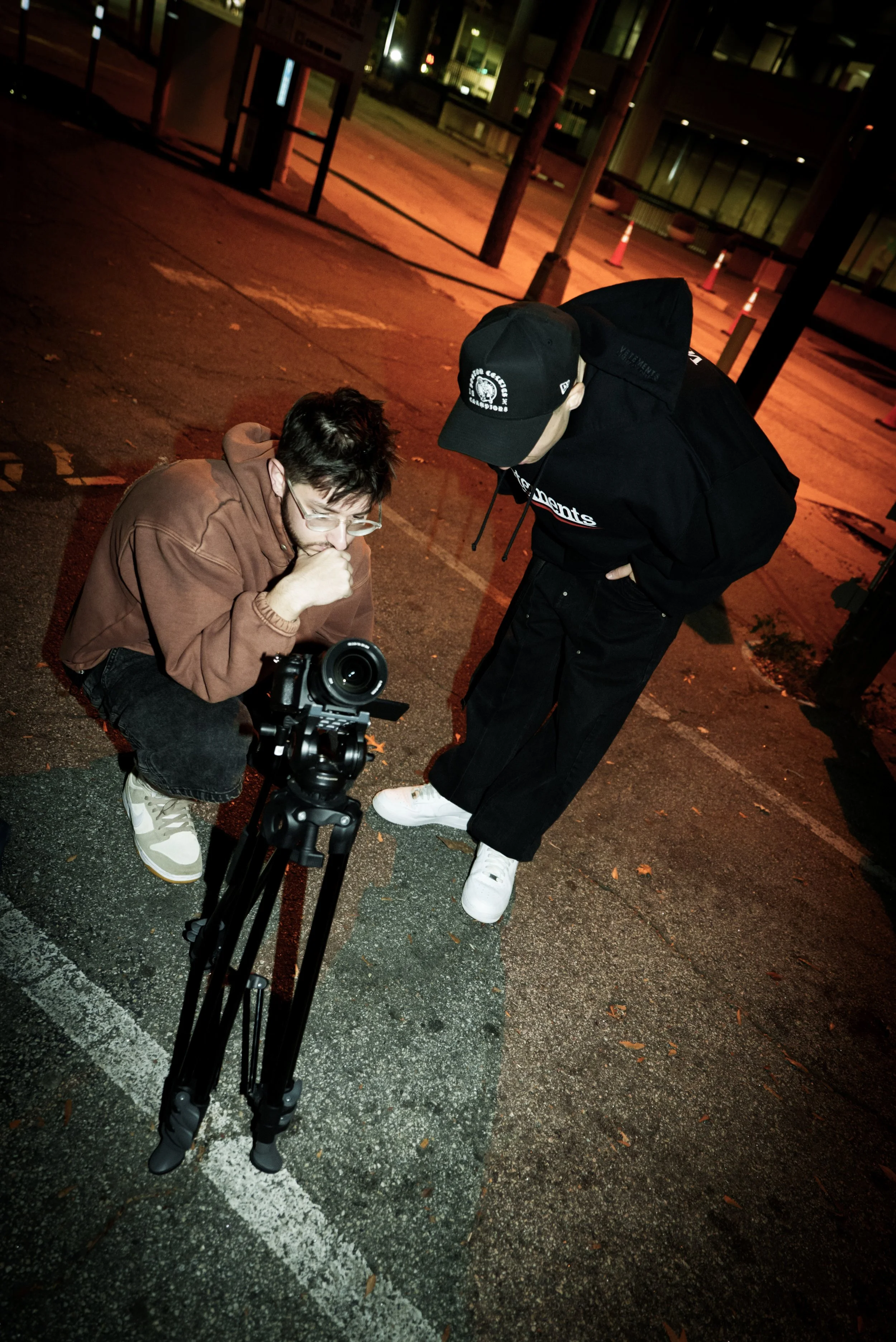 Token the rapper and nic violets at night, one squatting, looking at a camera on tripod, the other standing, observing, in an urban area with orange street lighting.
