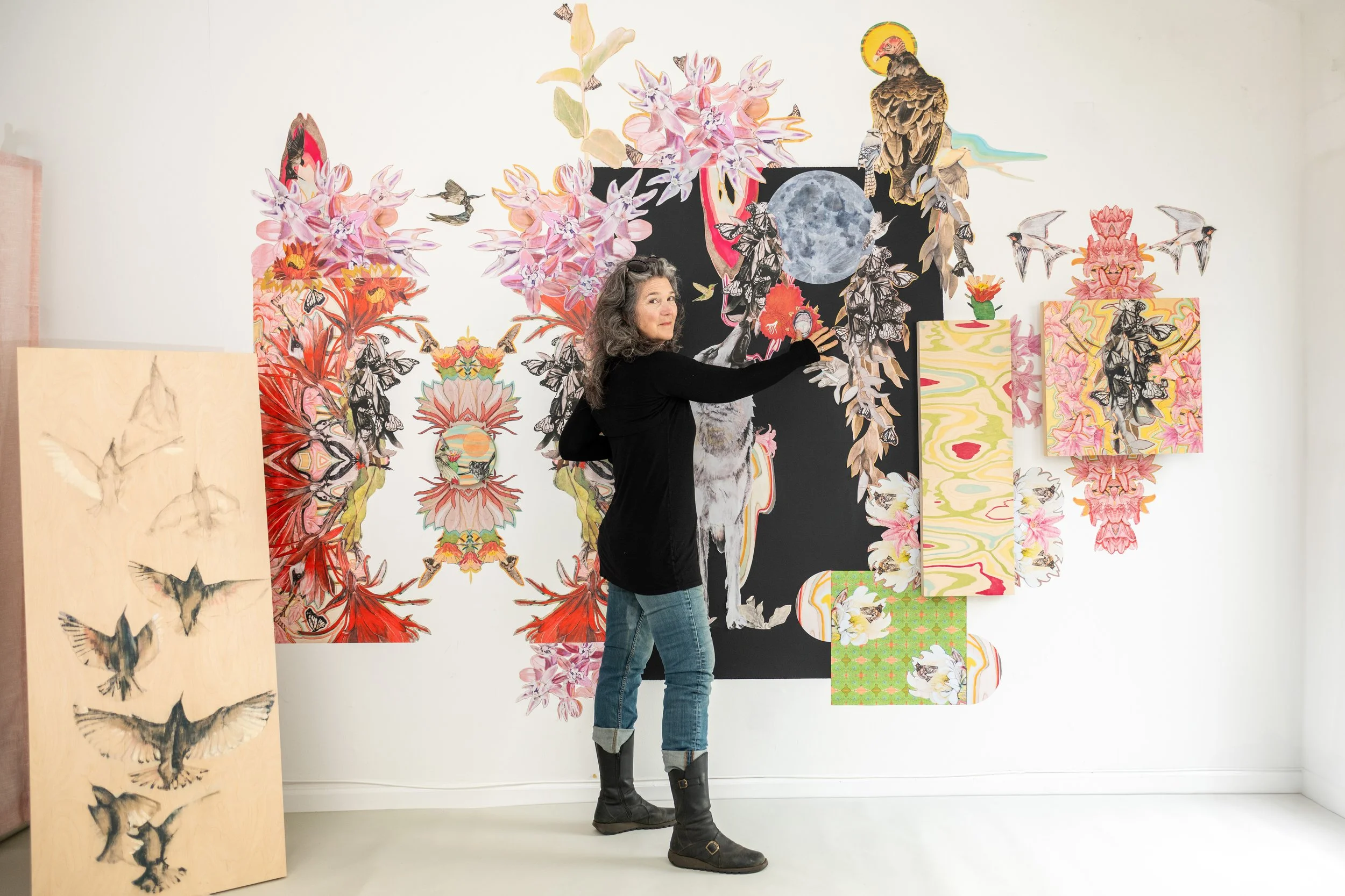 Portrait of artist standing partially facing artwork and looking back at camera. Art work, drawings, collage, paintings cover the wall while artist looks at camera.