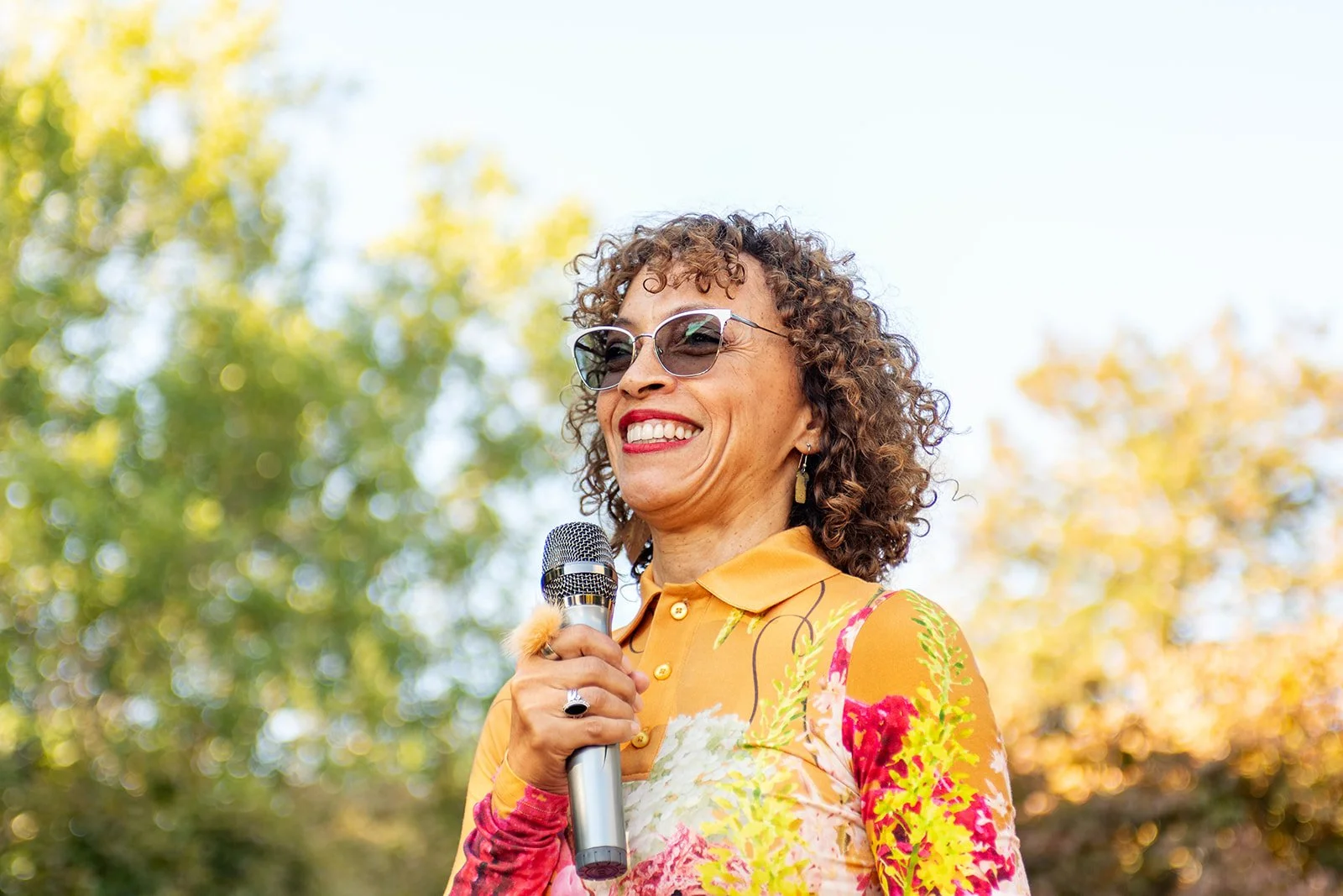 A woman with curly hair and sunglasses smiling while holding a microphone outdoors during daytime, with trees and clear sky in the background.
