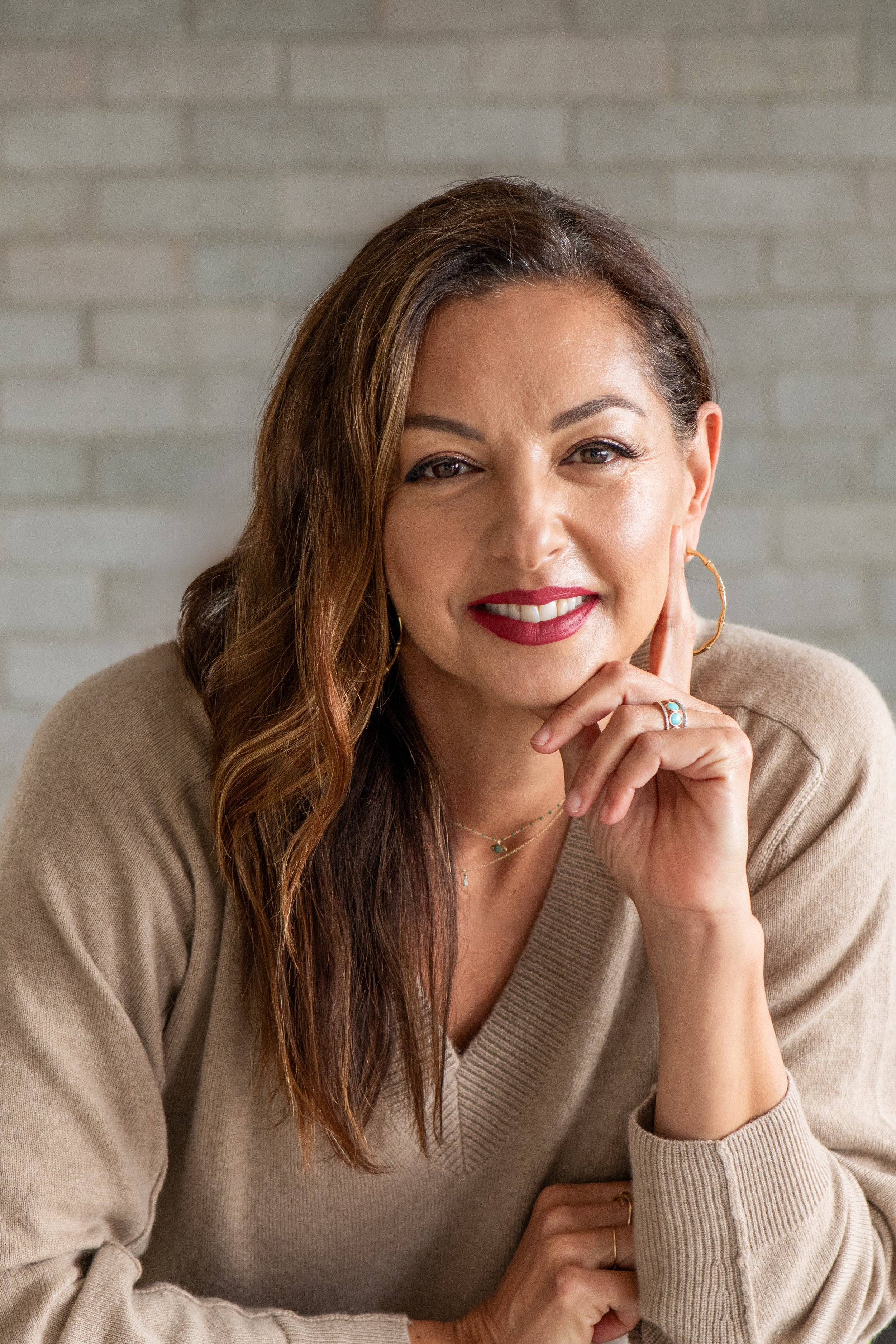 A woman with long brown hair, wearing a beige sweater, smiling, and resting her chin on her hand with jewelry and hoop earrings, in front of a light-colored brick wall.