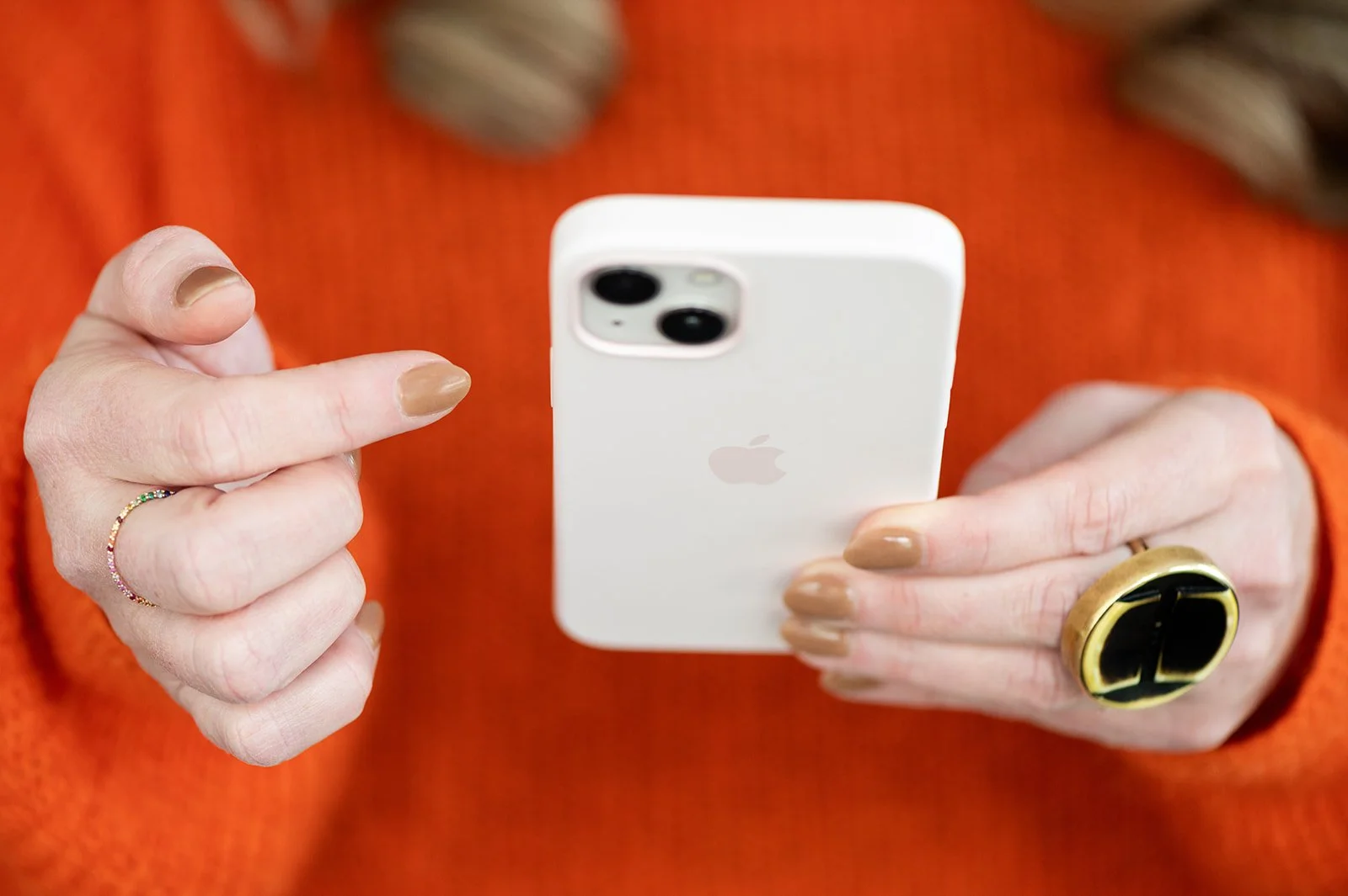Person holding a white iPhone with a camera module, wearing a large gold and black ring on their right hand, a small rainbow-colored ring on their left hand, and an orange fabric shirt in the background.