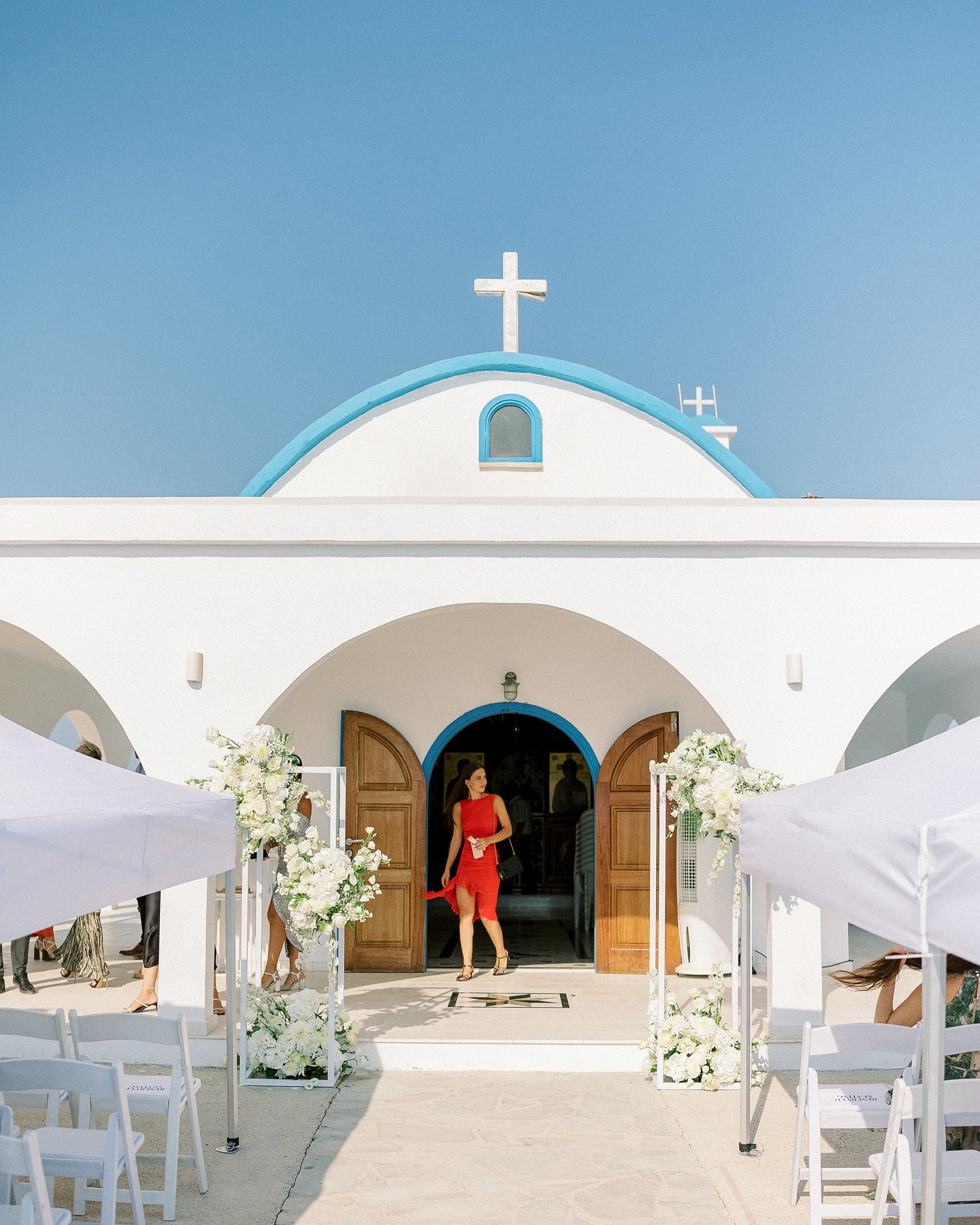 No plans, no poses, just contrast in perfect harmony.
A woman in a flowing red dress,
gliding past a chapel dressed in white and blue,
like fire meeting the sky.