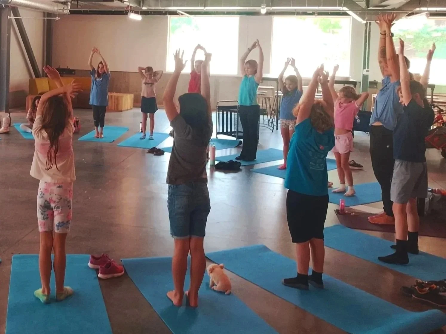 A group of young children participating in a yoga class, sitting or lying on purple yoga mats in a semicircular formation, led by an instructor inside a brightly decorated room.