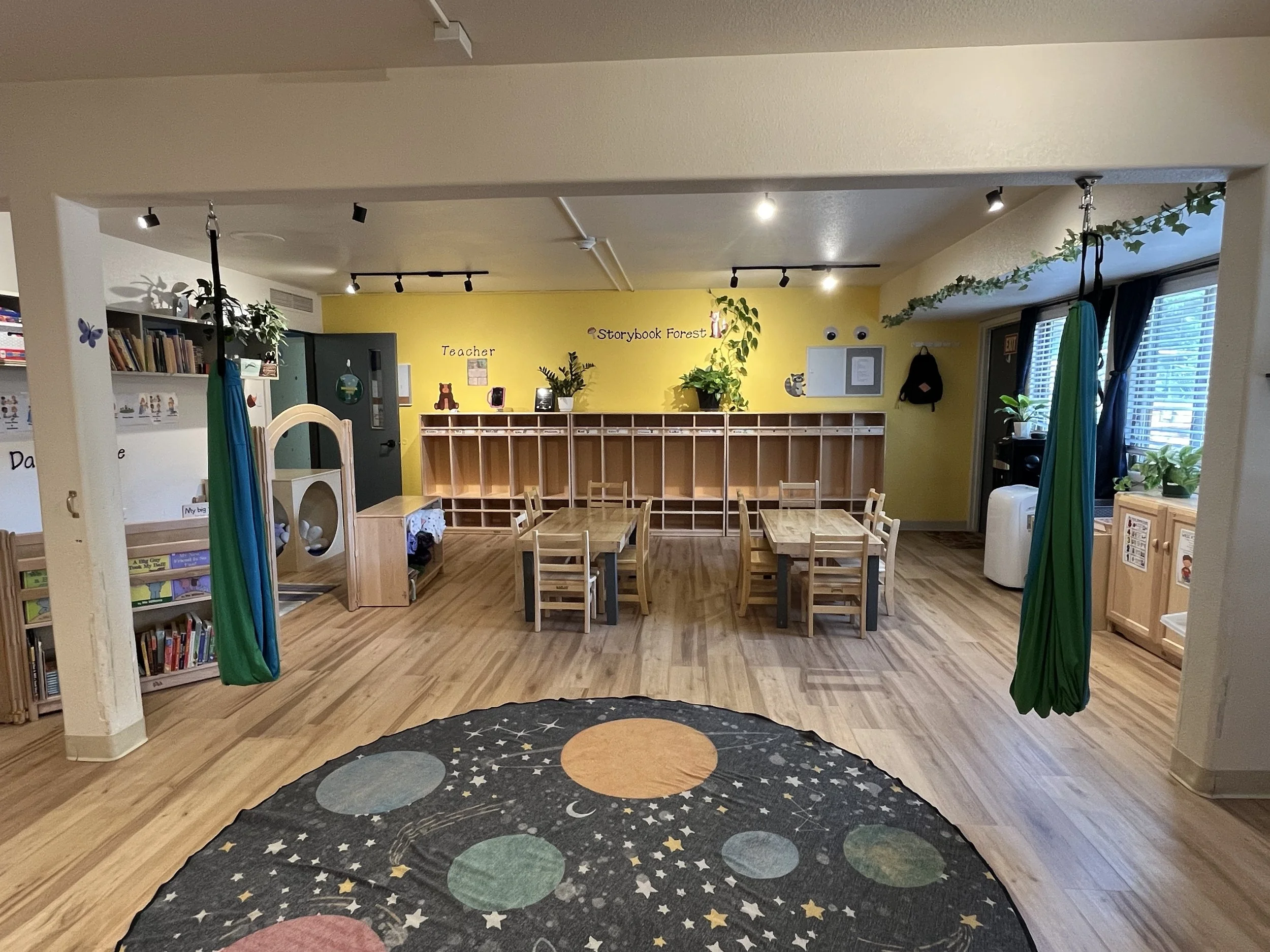 A classroom with small wooden tables and chairs, colorful decor, bookshelves, leaf decorations, and large windows with plants, indicating it's a preschool or kindergarten setting.