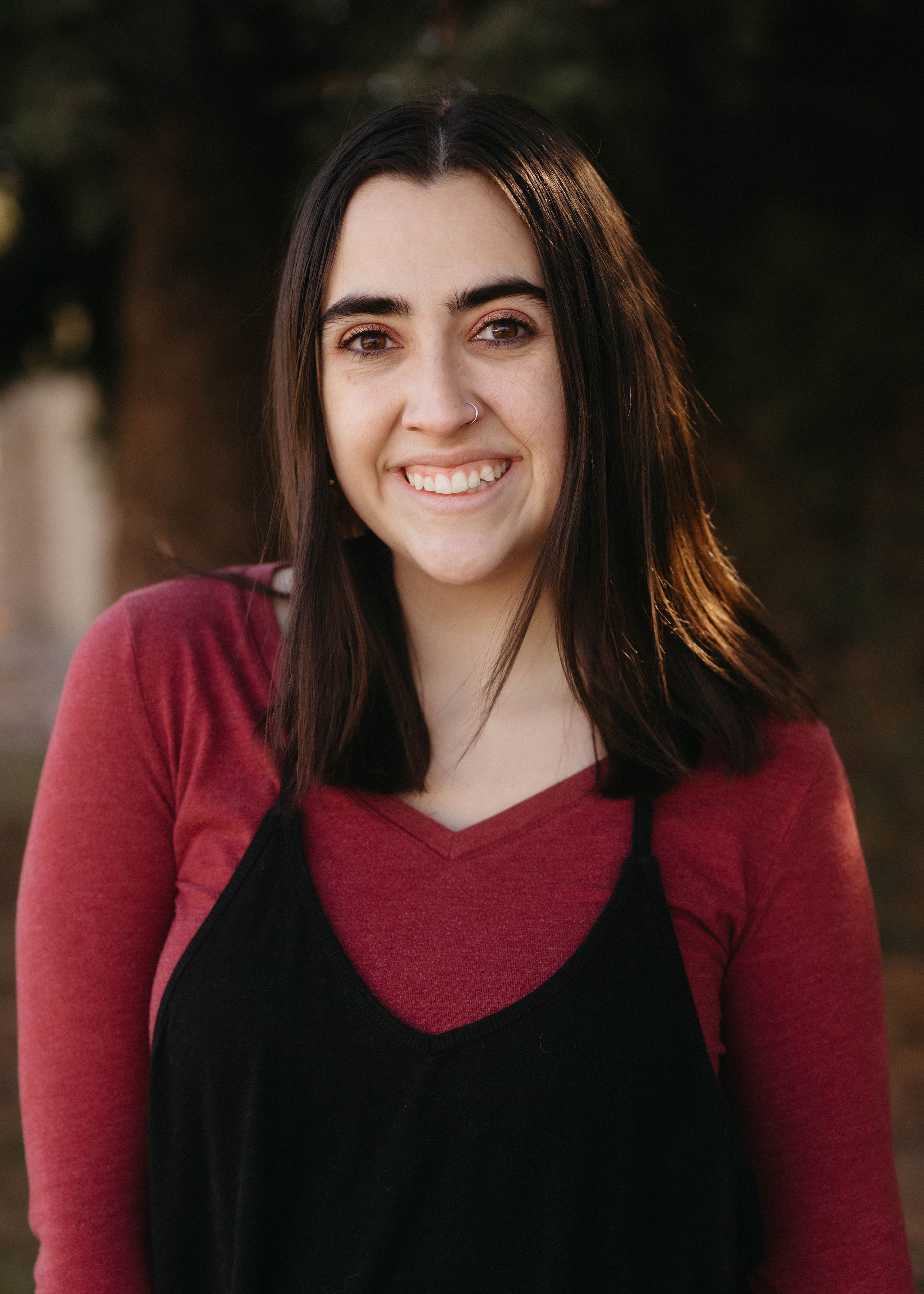 A young woman with straight dark hair and light skin, smiling in outdoor setting during golden hour, wearing a red long sleeve shirt underneath a black dress.