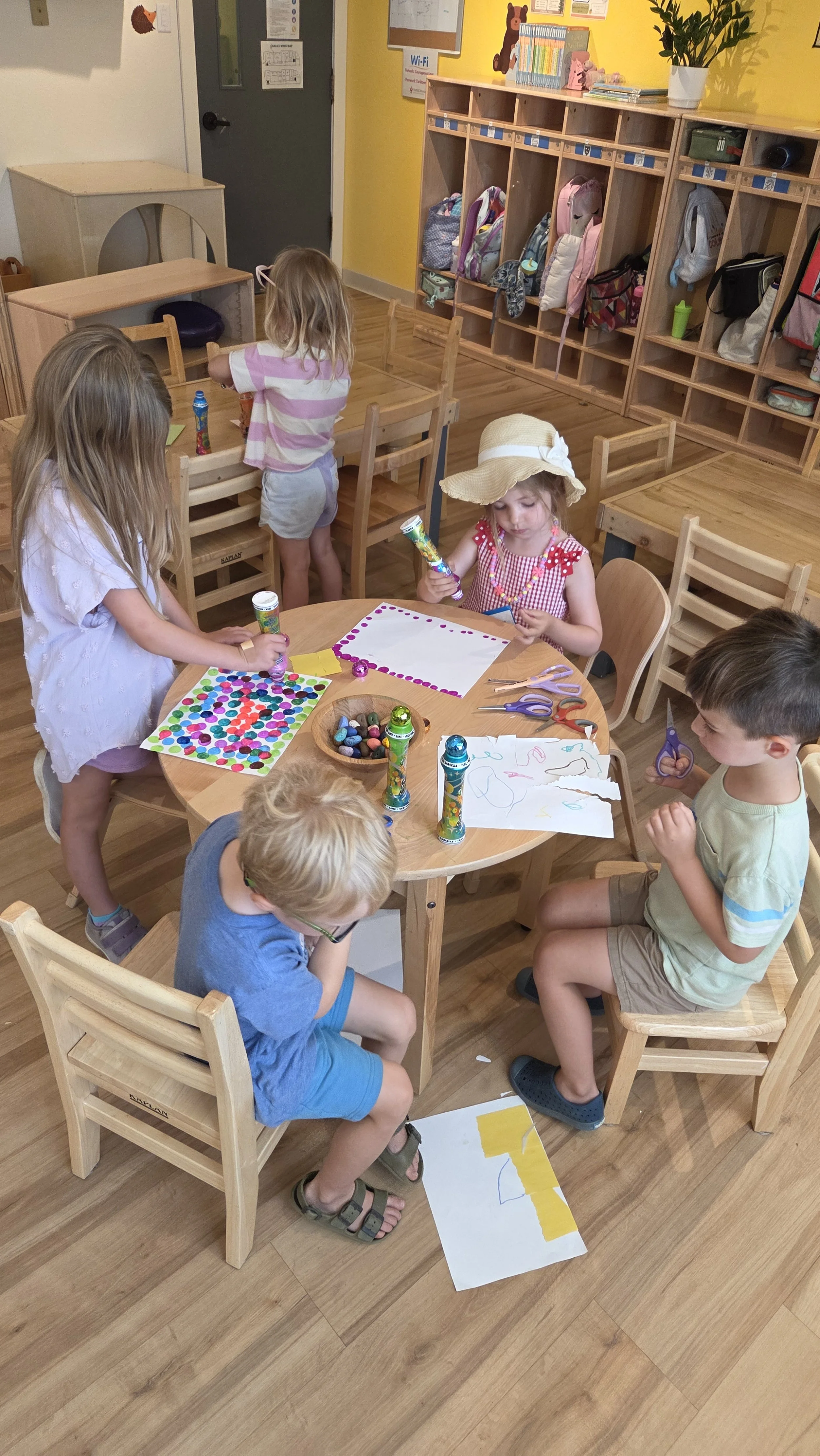 A group of children engaged in arts and crafts activities in a classroom. They are using glue, scissors, colored paper, and markers at a round wooden table.