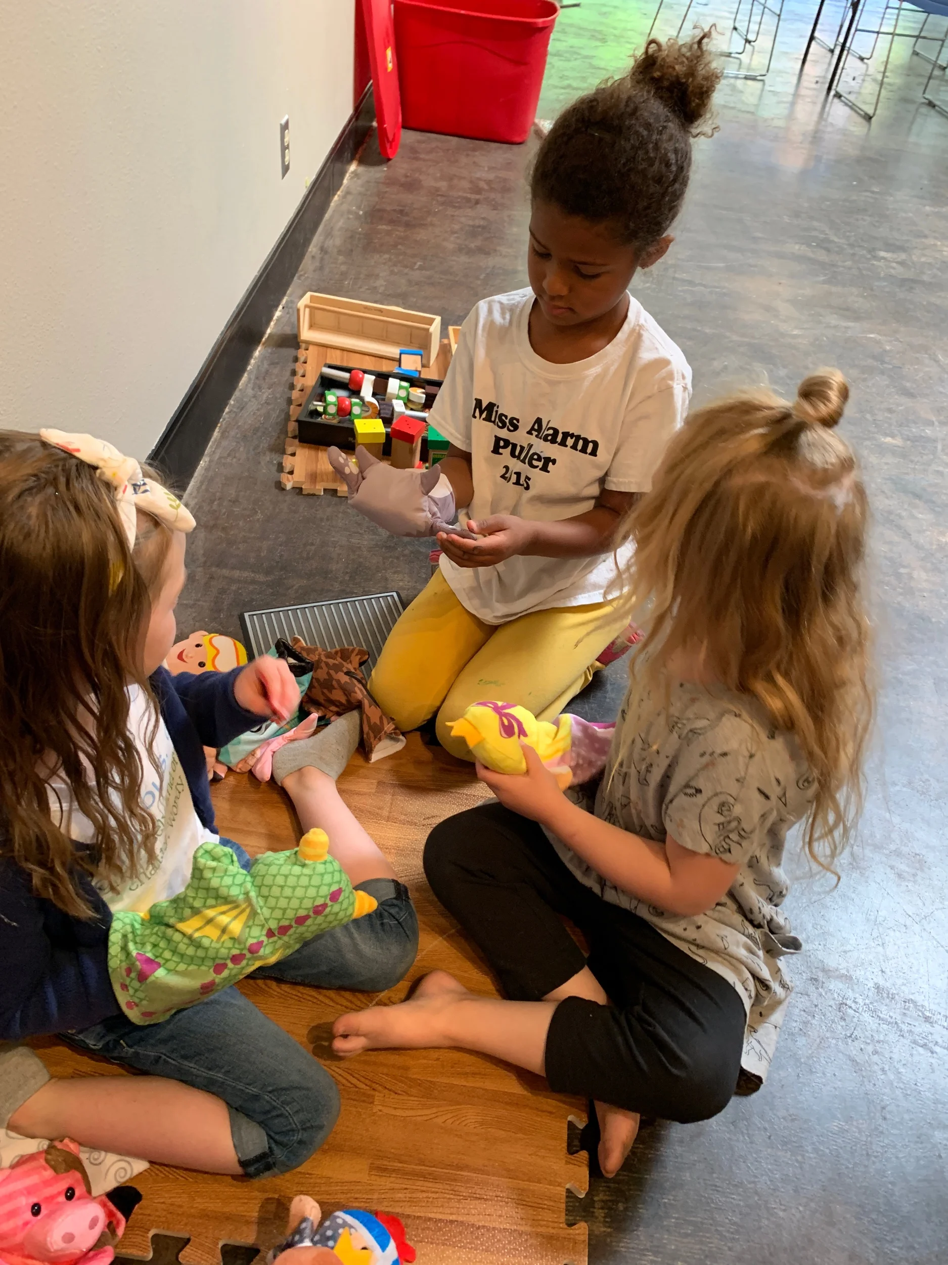 Three young girls playing with stuffed animals and toys on a wooden floor, with a box of toys behind them and a woman kneeling in front of them.