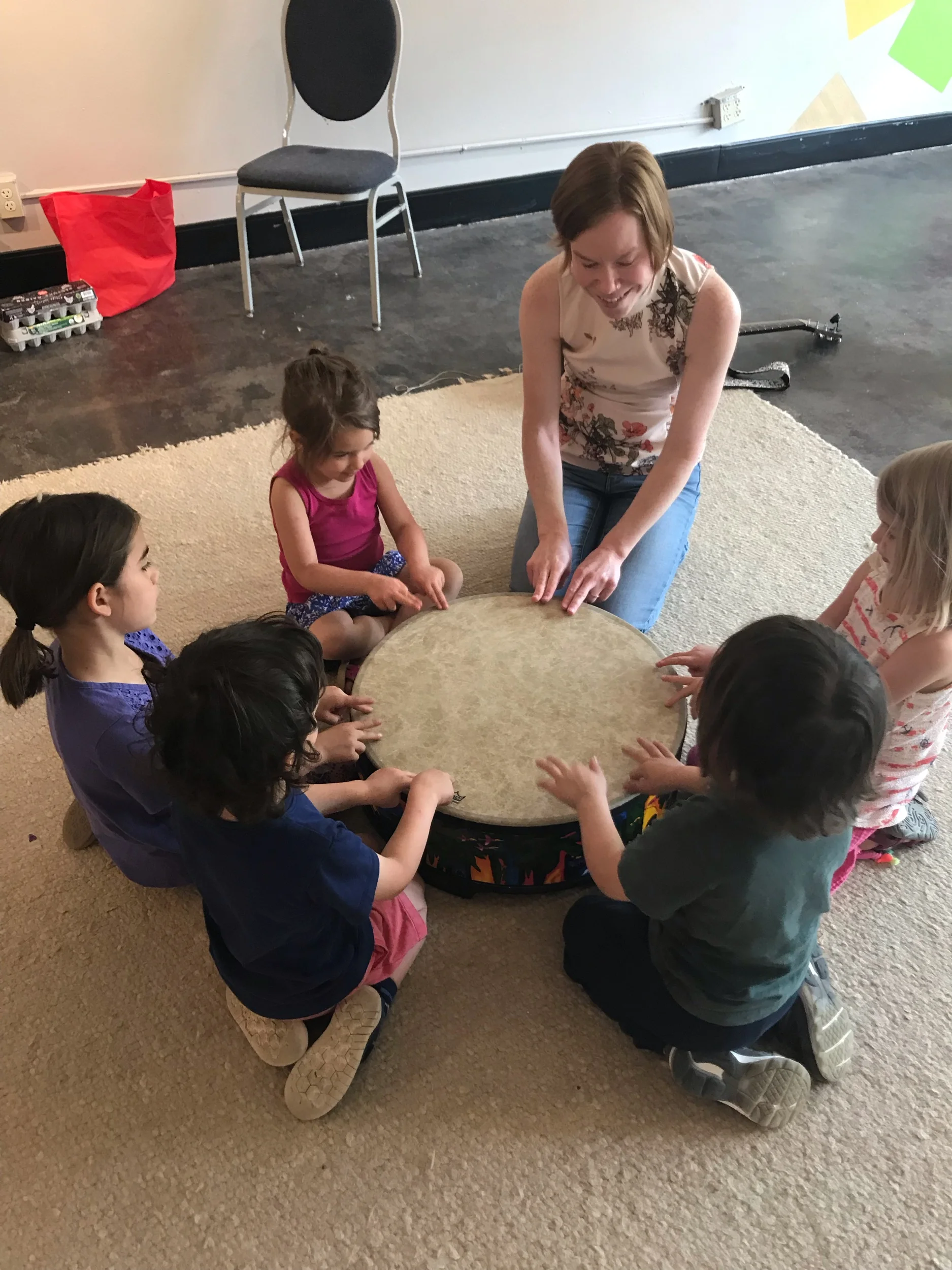 A group of five children sitting cross-legged around a drum on a carpeted floor, with a woman kneeling and leading them in a musical activity in a room with a black chair, a red bag, and some supplies in the background.