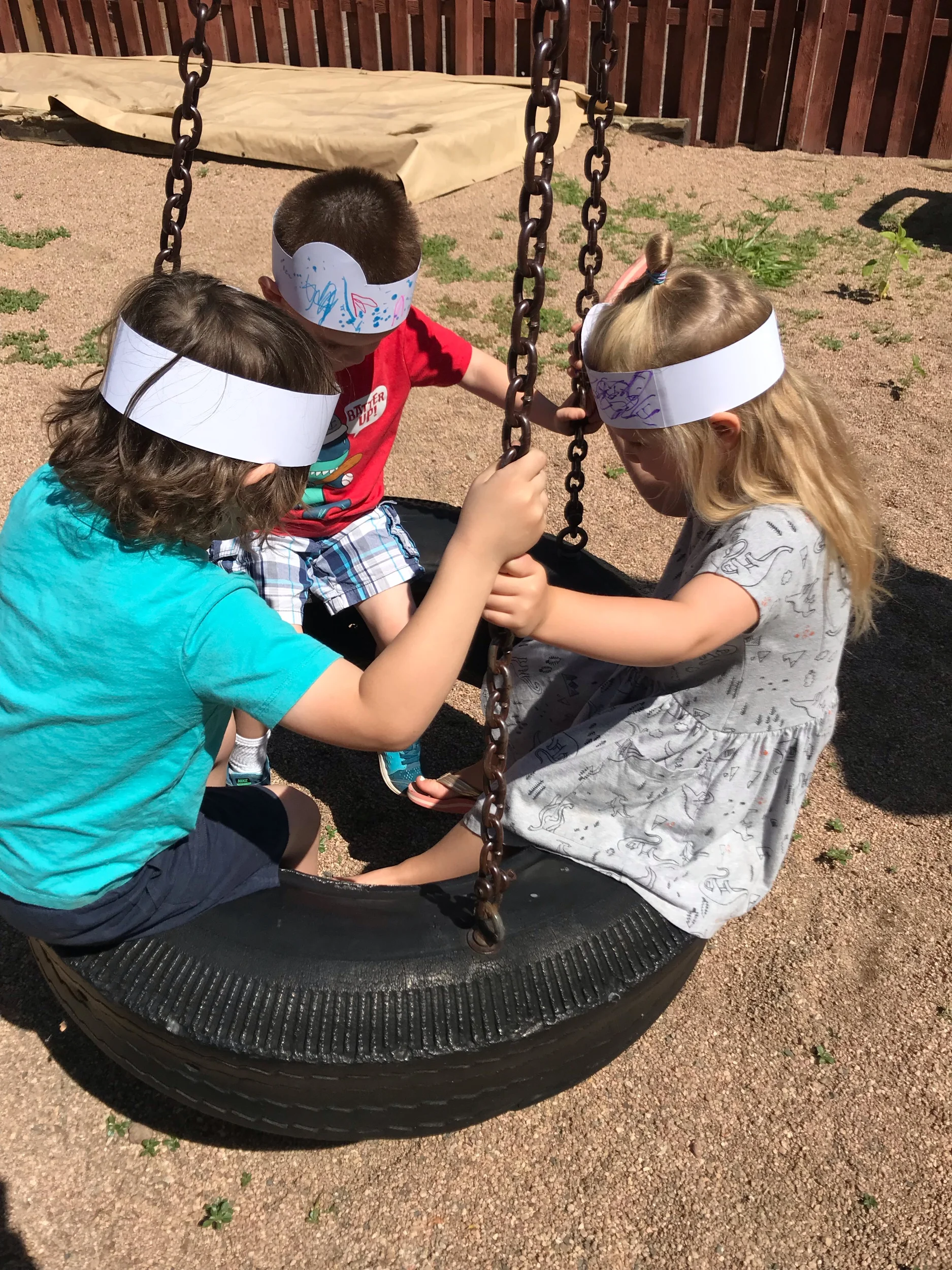 Three young children sitting inside a tire swing, holding onto the chains, and wearing paper headbands with drawings, at a playground on a sunny day.