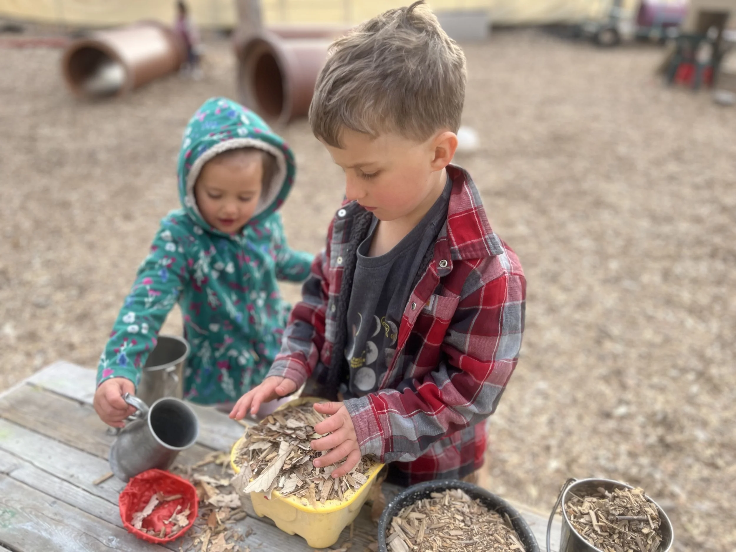 Two children, a boy and a girl, playing outdoors with wood chips and small metal buckets on a wooden table in a playground.