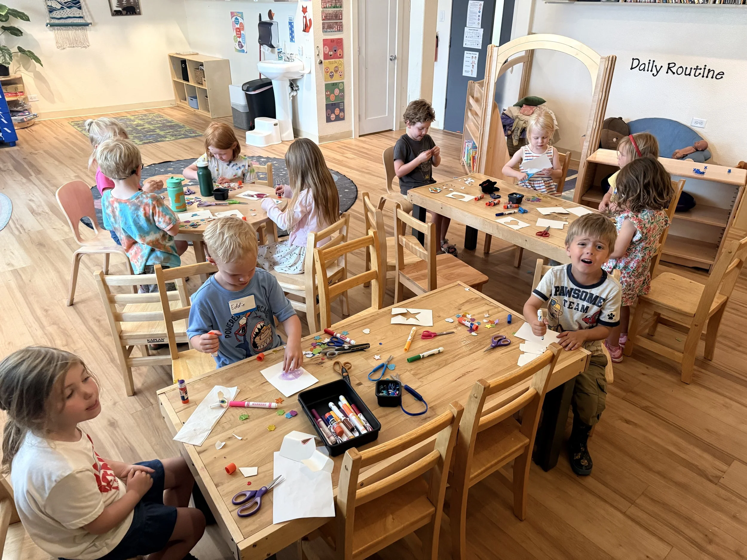 Young children engaged in arts and crafts activities at wooden tables in a classroom, with art supplies like glue, scissors, and paper scattered around.