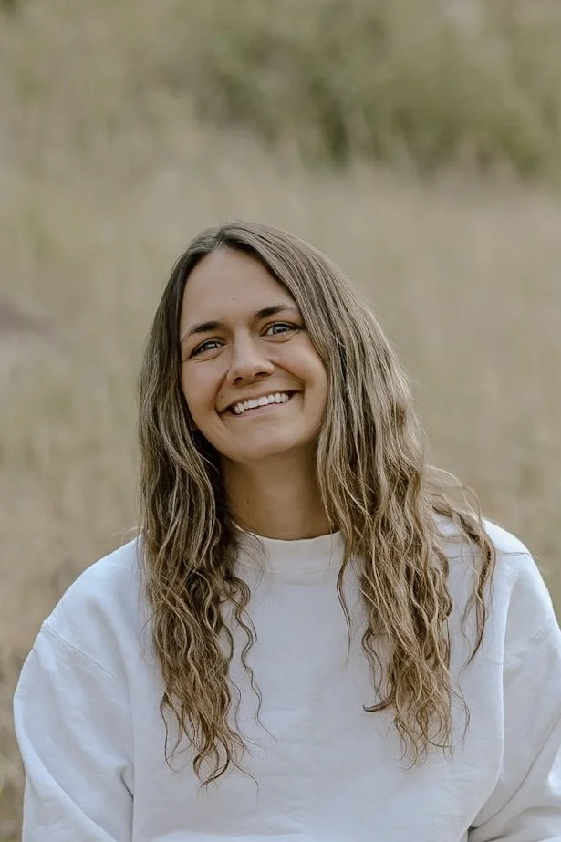A woman with long wavy brown hair smiling outdoors with a blurred grassy background.