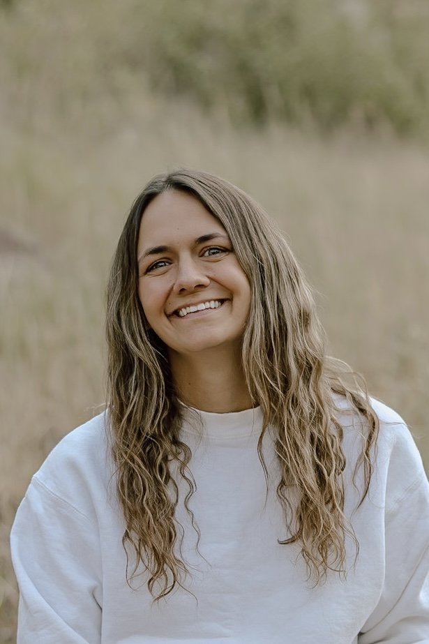 A woman with long wavy brown hair smiling outdoors with a blurred grassy background.