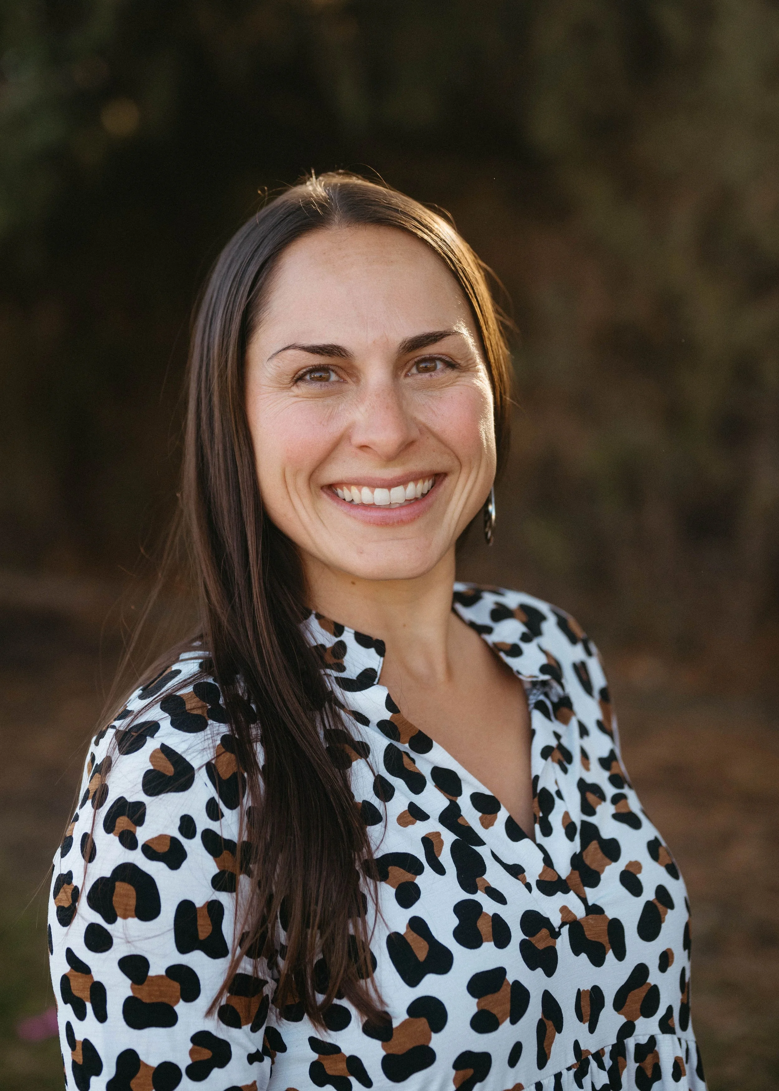 A smiling woman with long dark hair, wearing a white blouse with a black and brown animal print pattern, outdoors with blurred natural background.