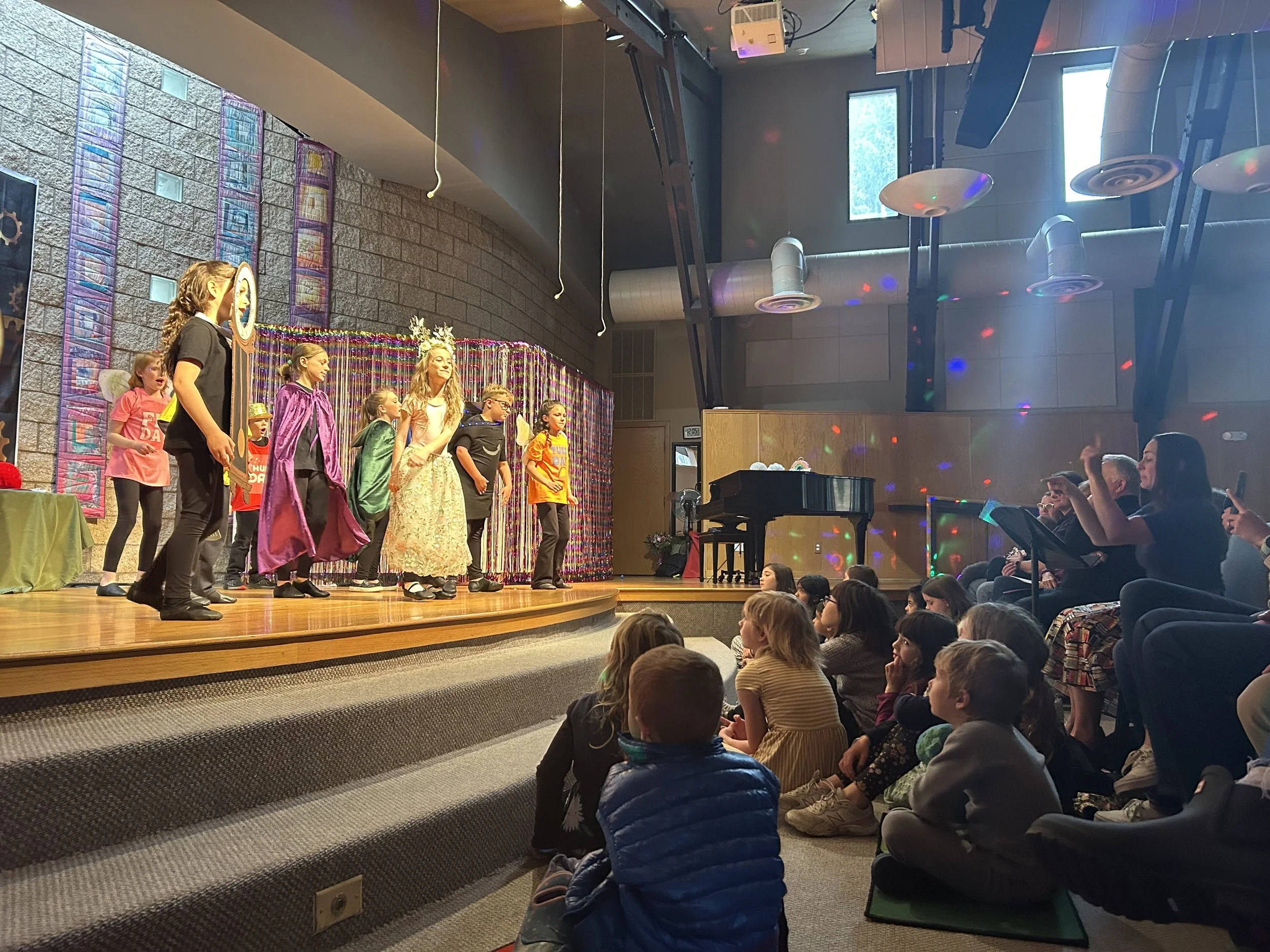 Children performing on stage in costumes with an audience of children and adults watching indoors at a school or community center