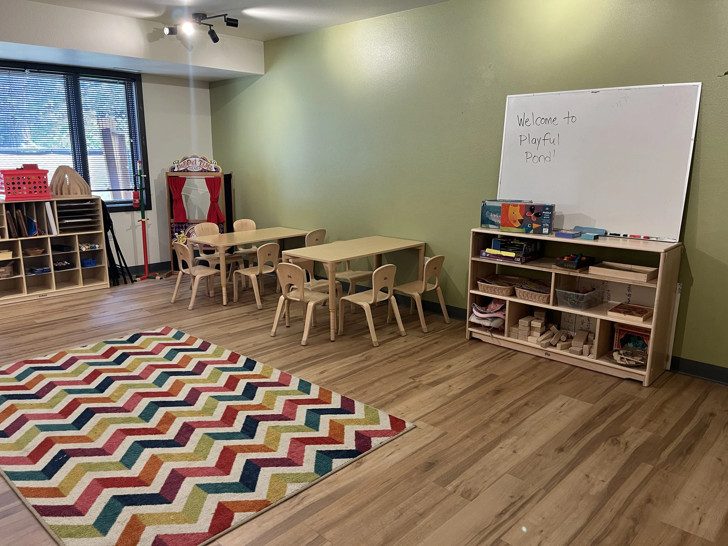 A preschool classroom with small wooden tables and chairs, a colorful zigzag patterned rug, a whiteboard with writing, and shelves filled with educational toys and supplies.