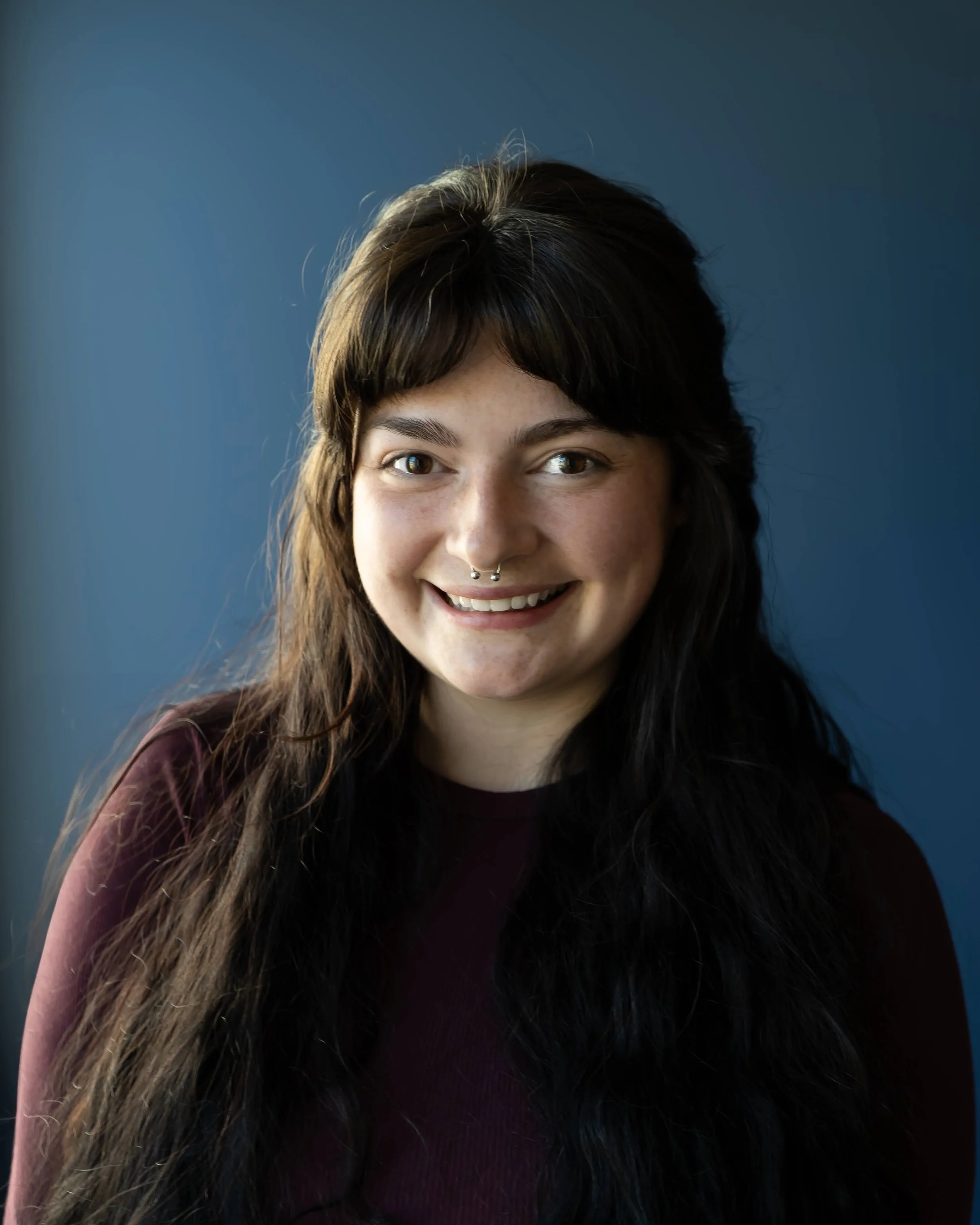 A young woman with long, dark hair, a septum piercing, and wearing a maroon top, smiling, set against a solid blue background.