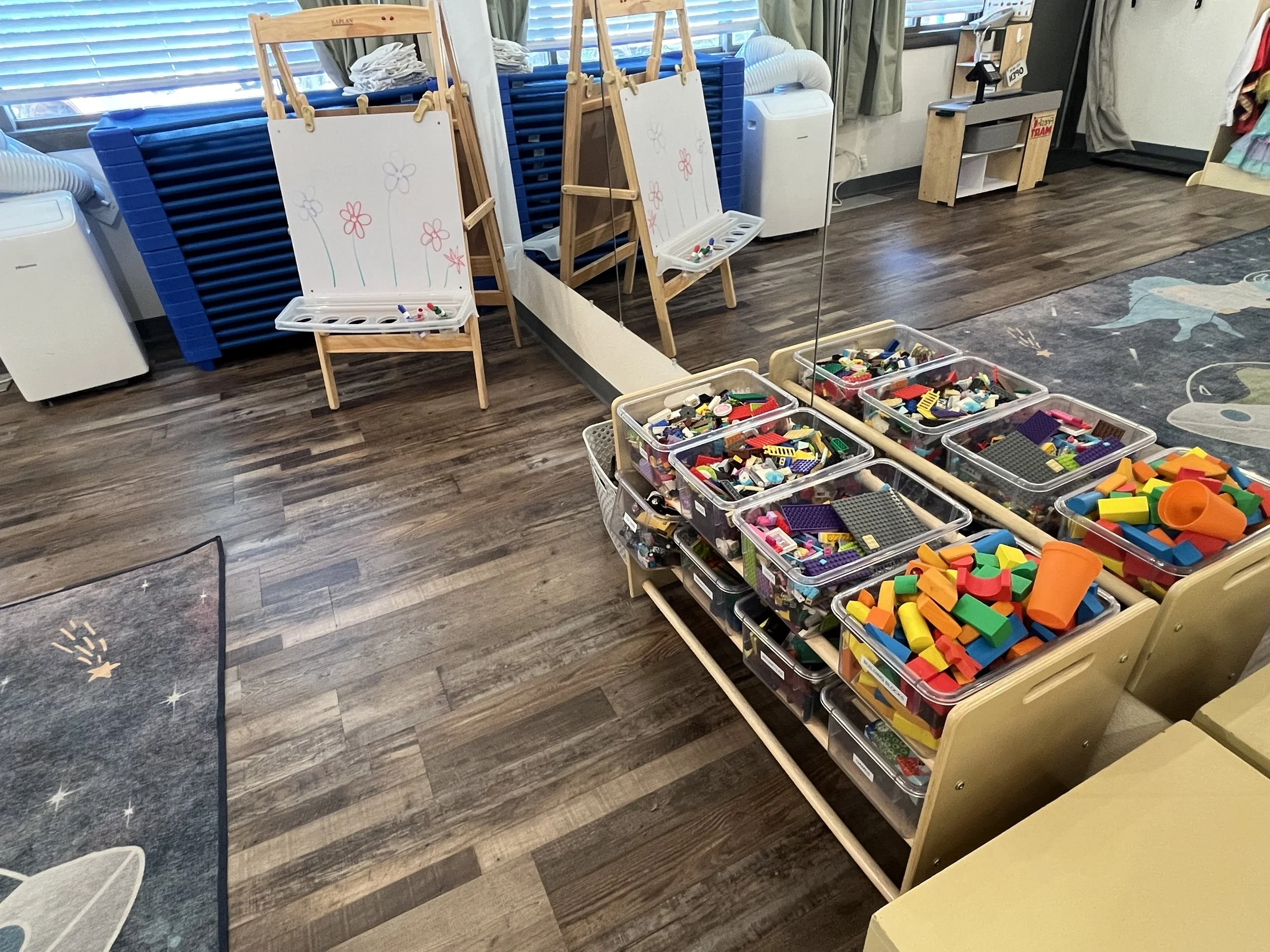A play area with large clear plastic bins filled with colorful building blocks and LEGO pieces, and two small wooden easels with whiteboards decorated with simple flower drawings, set in a room with wooden flooring and a mirror, near a window with blinds.