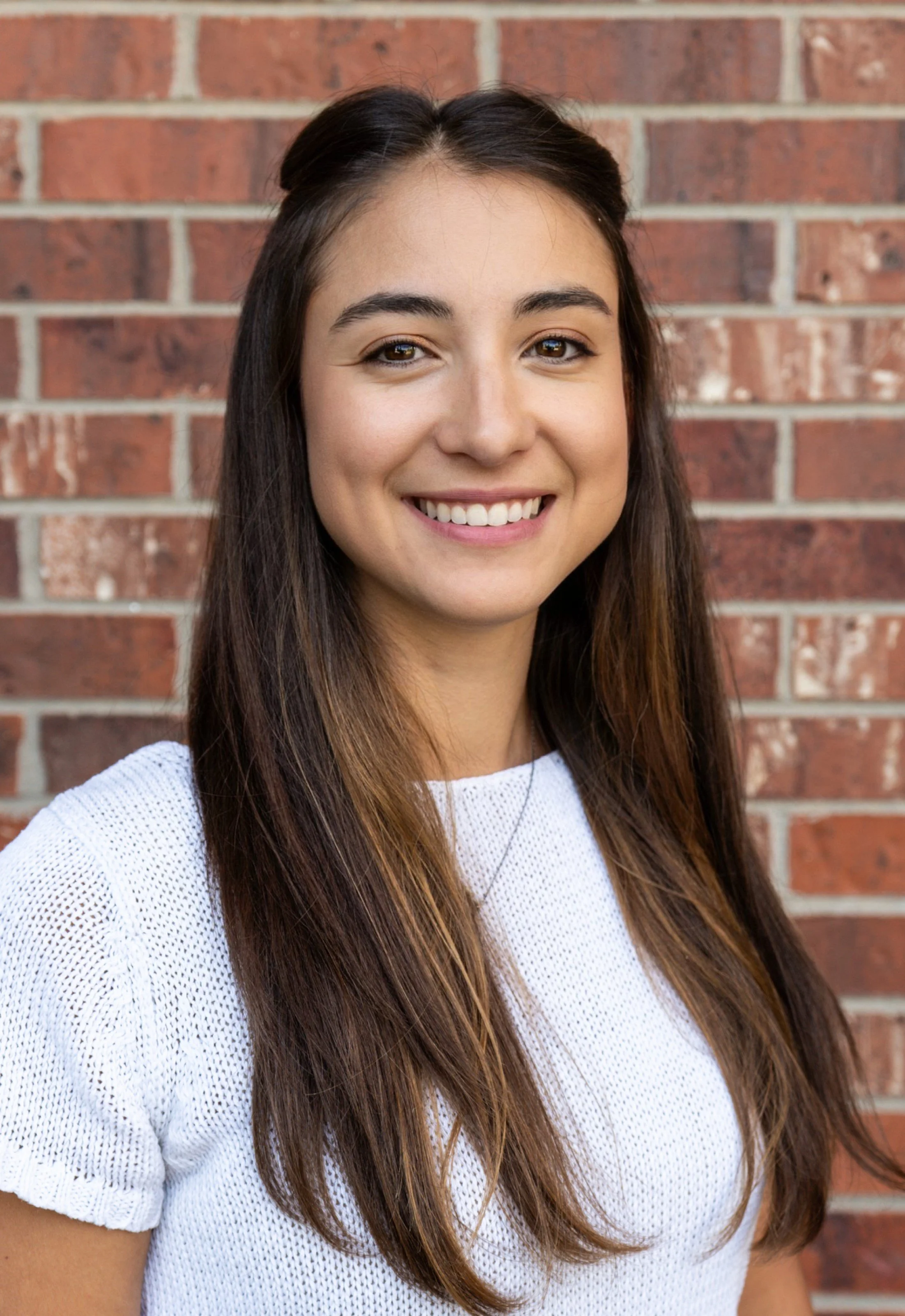 A young woman with long brown hair and brown eyes smiling in front of a brick wall, wearing a white knit top.