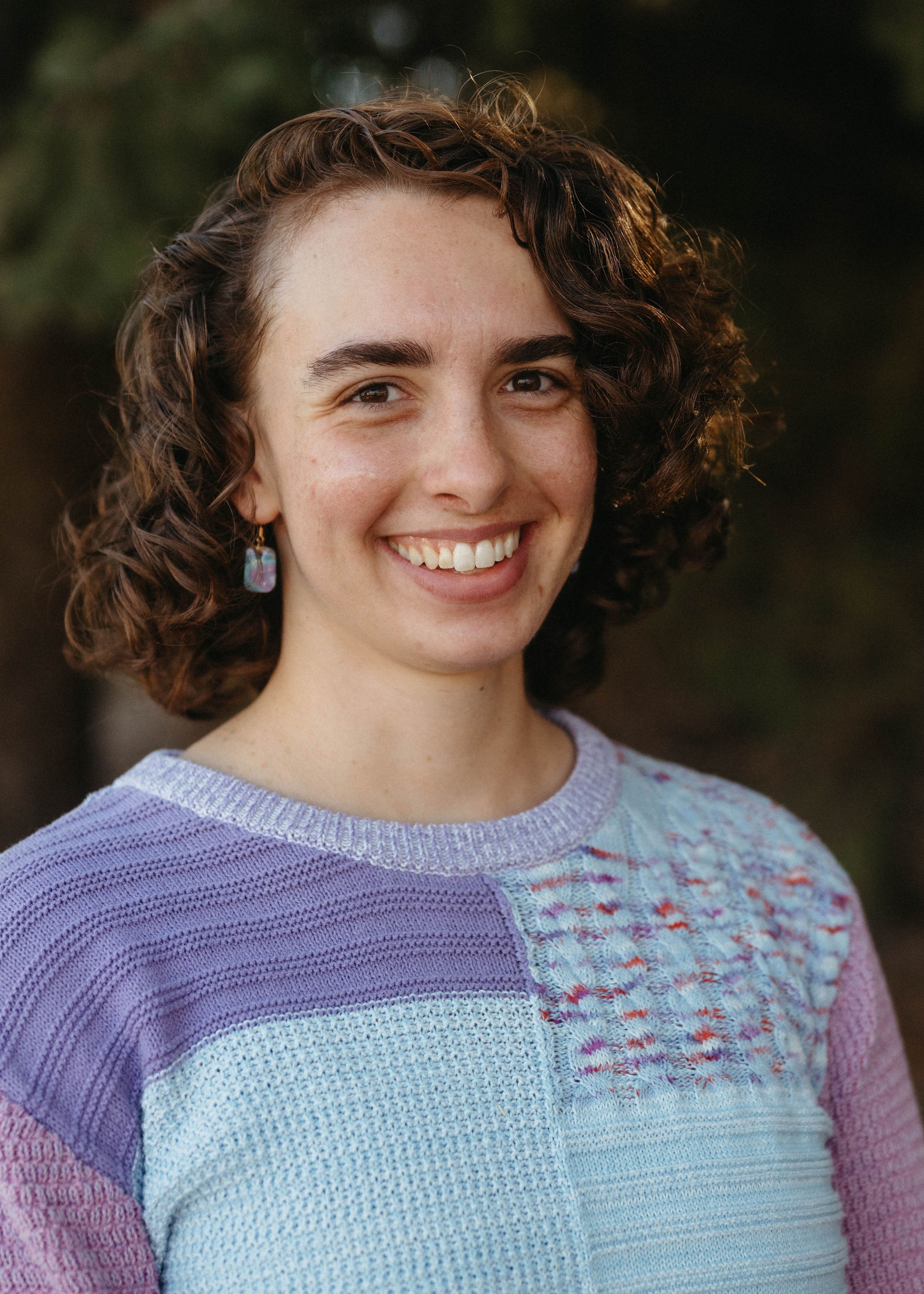 A young woman with curly brown hair, smiling outdoors, wearing a multicolored knitted sweater and colorful earrings.