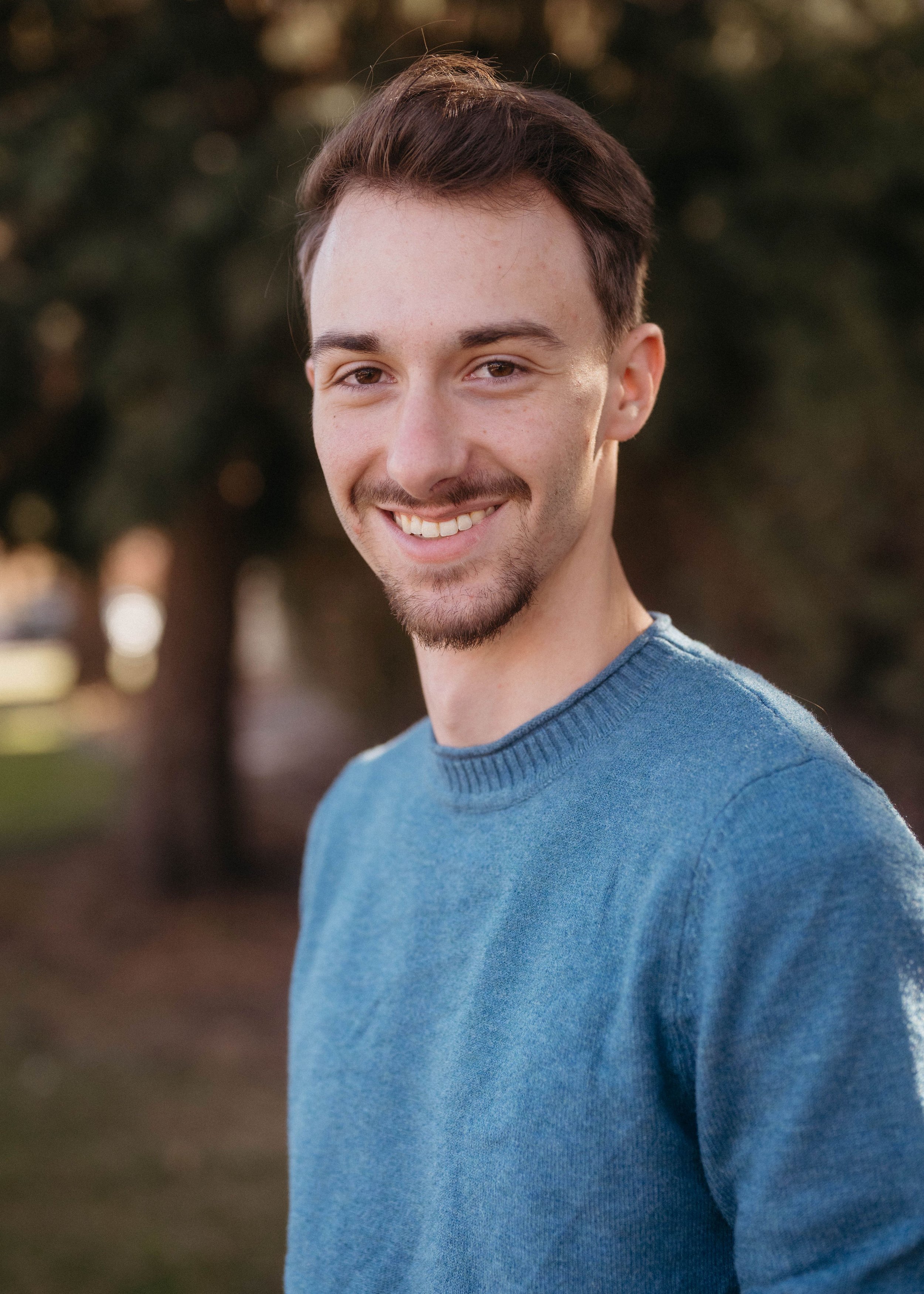 A young man with short brown hair, a mustache, and a beard, smiling outdoors in a park during daytime, wearing a blue sweater.