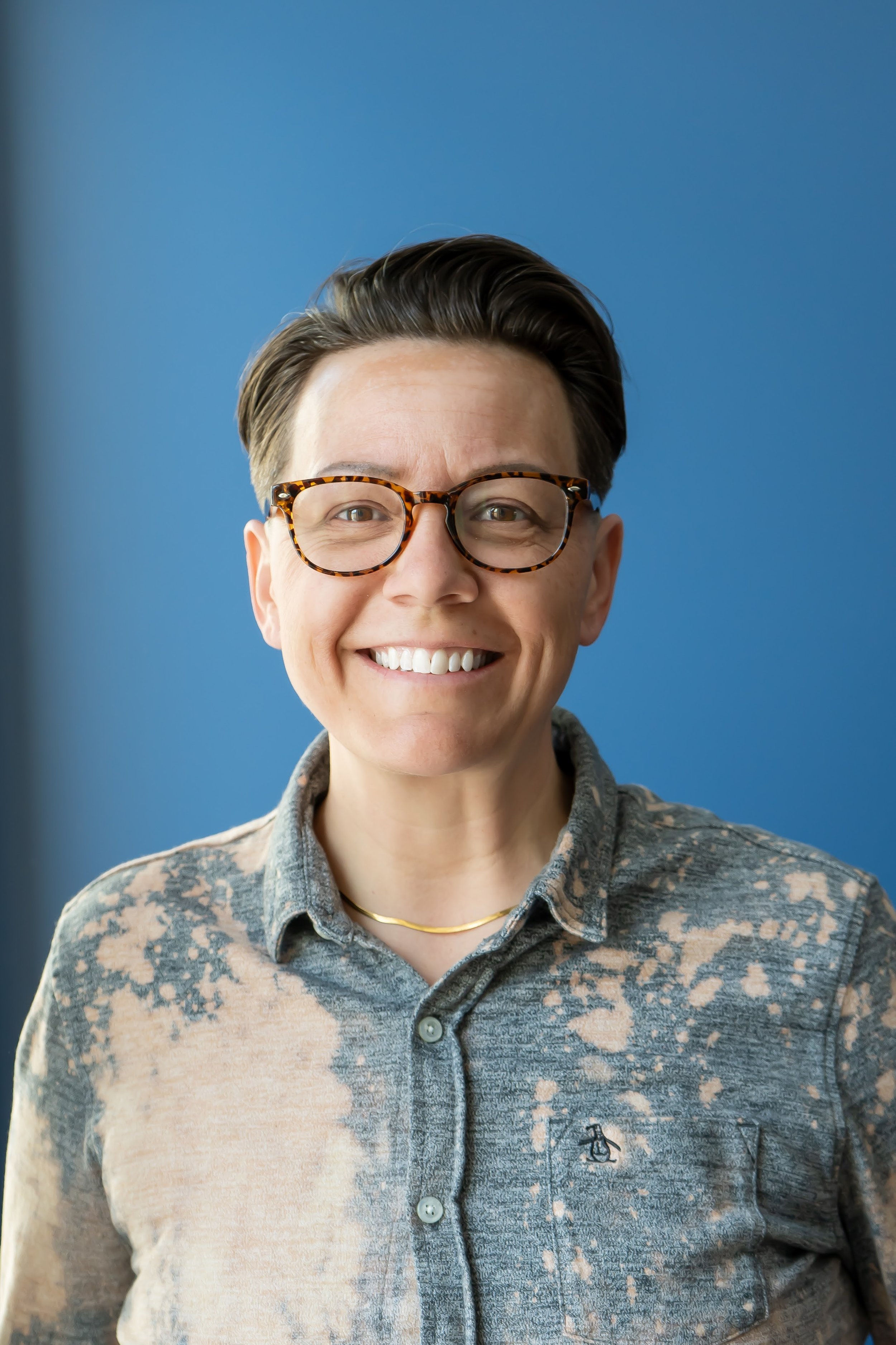 A woman with short dark hair, wearing glasses with a tortoise shell pattern, a patterned button-up shirt, and a gold necklace, smiling in front of a blue background.