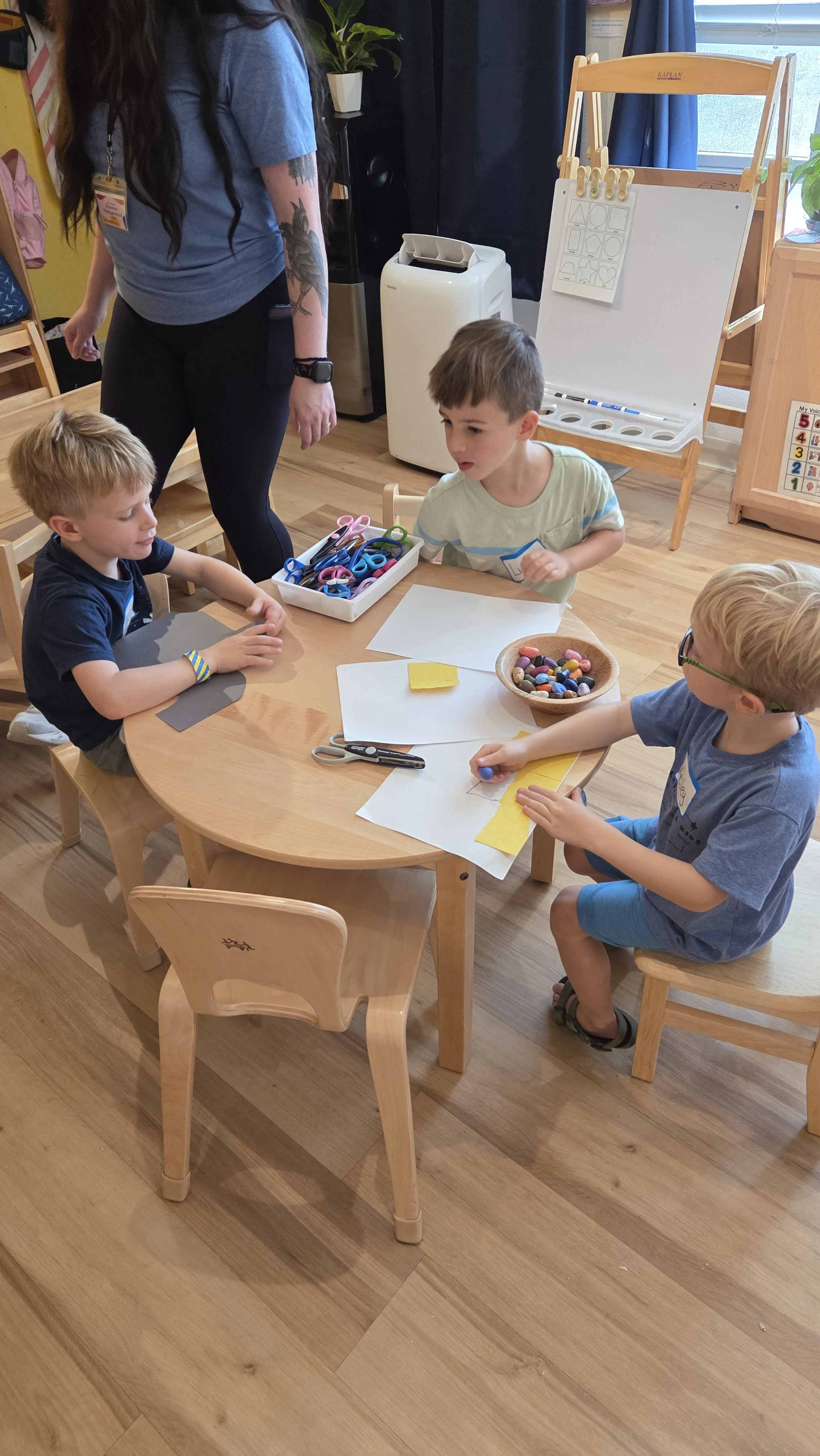 Three young boys sitting at a wooden table doing arts and crafts with paper, scissors, and colored objects, with a woman standing nearby overseeing the activity in a classroom