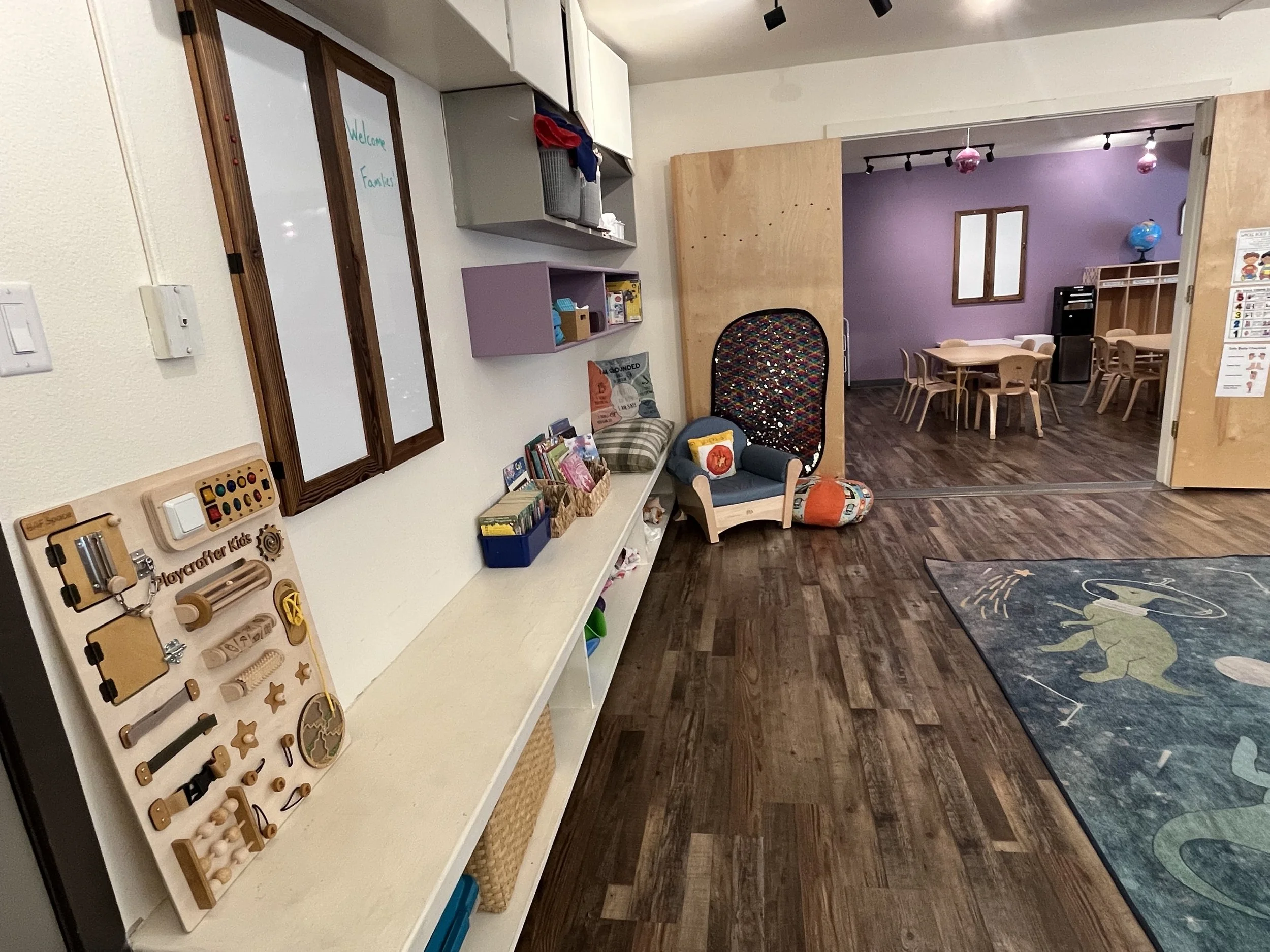 A view of a classroom or play area with wooden floors, a white tabletop with toys and books, a small blue and yellow armchair, and tables with chairs in a room with purple and lavender walls, decorated with globe and other educational items.