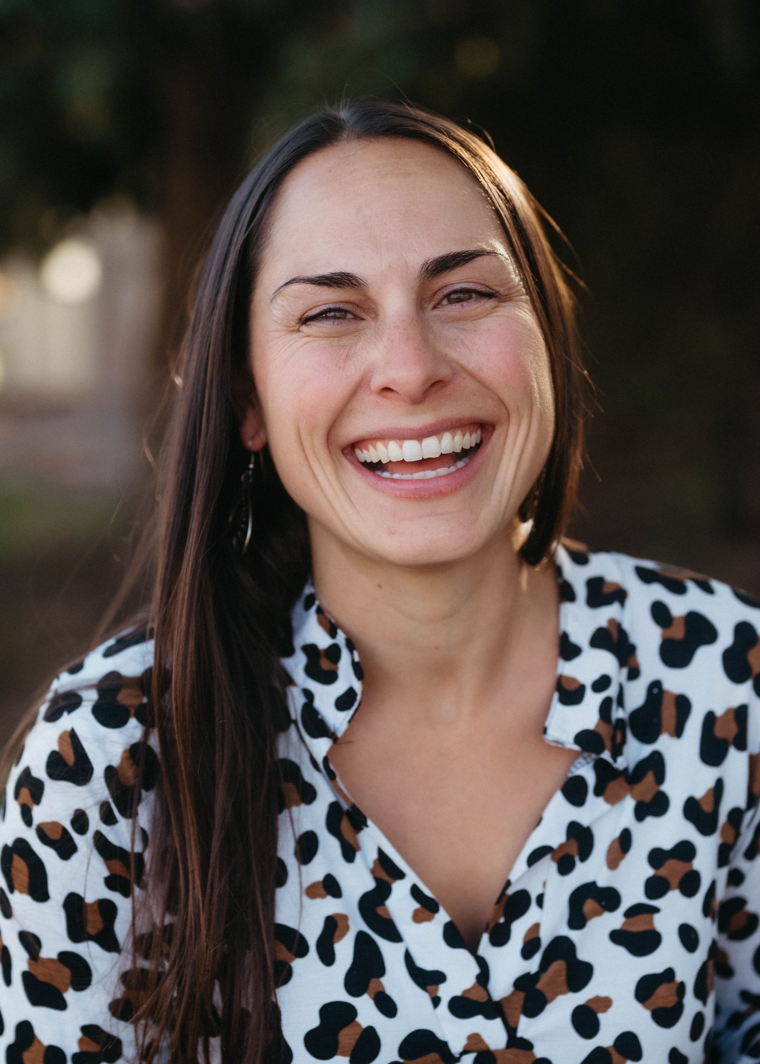 Close-up of a woman with long dark hair smiling outdoors, wearing a white blouse with a black and brown leopard print pattern.