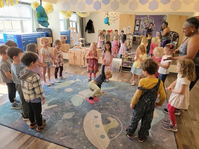 A woman reading a children's book titled "We Are In a Book!" to a group of children sitting and lying on a carpeted floor in a classroom or library.