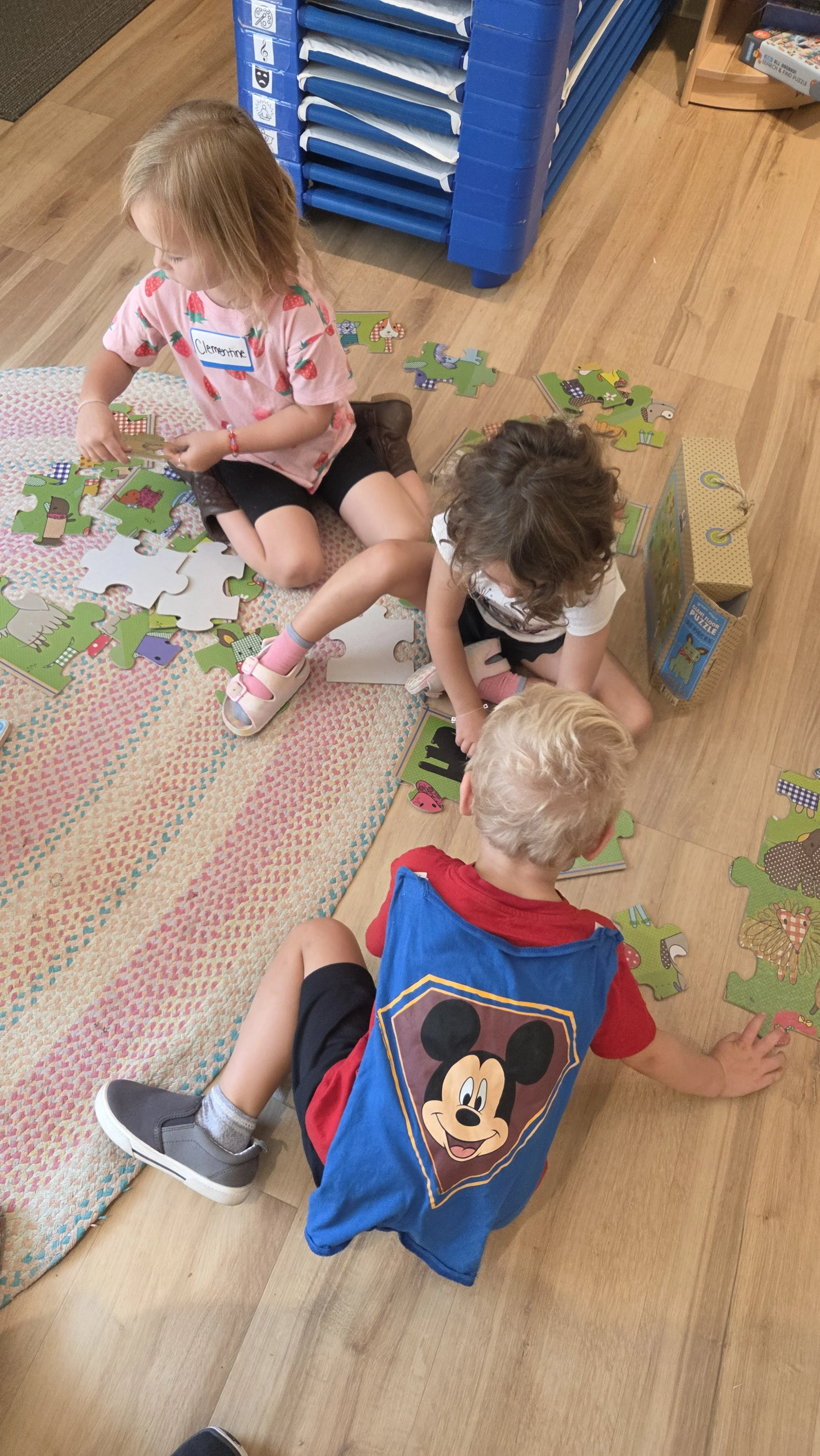 Three children are sitting on the wooden floor assembling a puzzle with farm animal images, with a blue storage rack and shelves nearby.